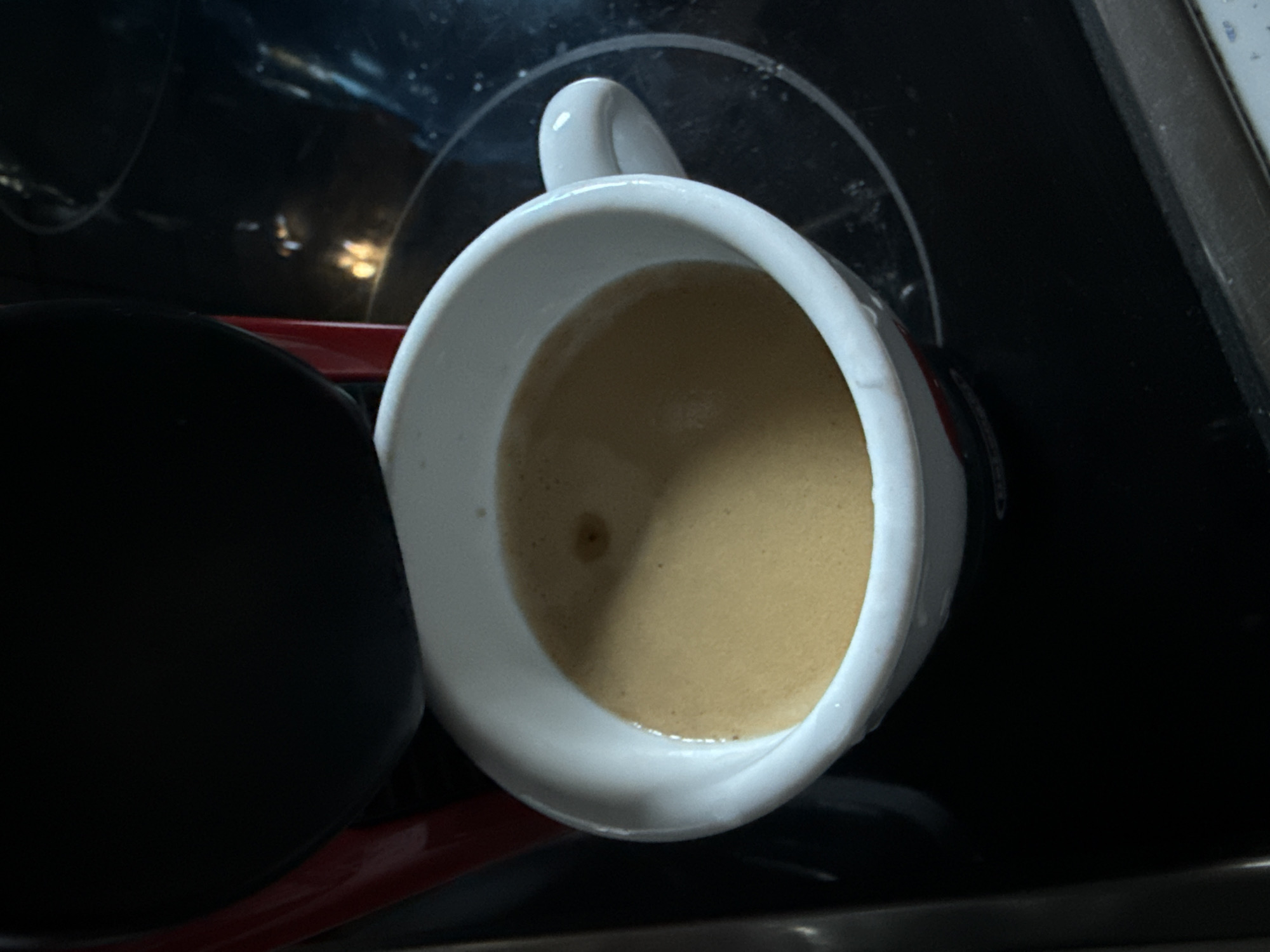 A close-up shot focuses on a white espresso cup filled with dark coffee, featuring a creamy crema layer. The cup rests on a black glass stovetop, which reflects some ambient light. The upper portion of the image is dominated by the dark, curved surface of a coffee machine, partially obscuring the scene. There are no people or animals visible in the image. The setting appears to be a kitchen in Wrocław, Poland.