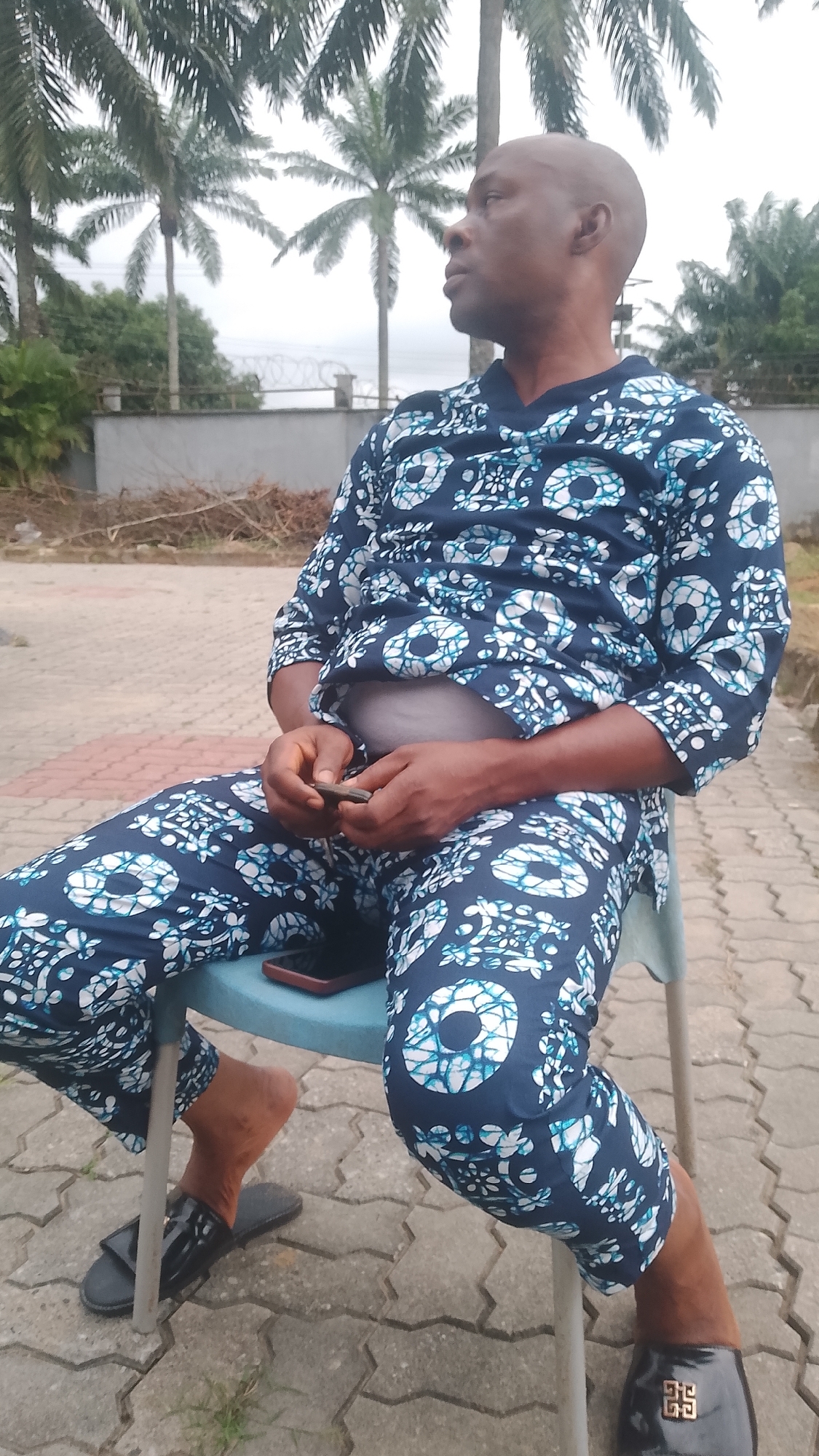 A man is sitting on a blue chair outdoors, looking to his right. He is wearing a navy blue dashiki with a white and blue circular pattern. His left hand is resting on his lap, holding a dark object, possibly a phone. A red phone is visible on the seat of the chair. He is also wearing black sandals. The setting appears to be paved with interlocking bricks. In the background, palm trees are visible against an overcast sky. A low concrete wall with a fence is also in the background. The time of day is daytime, and the weather appears to be cloudy. The location is Agbarho, Nigeria.