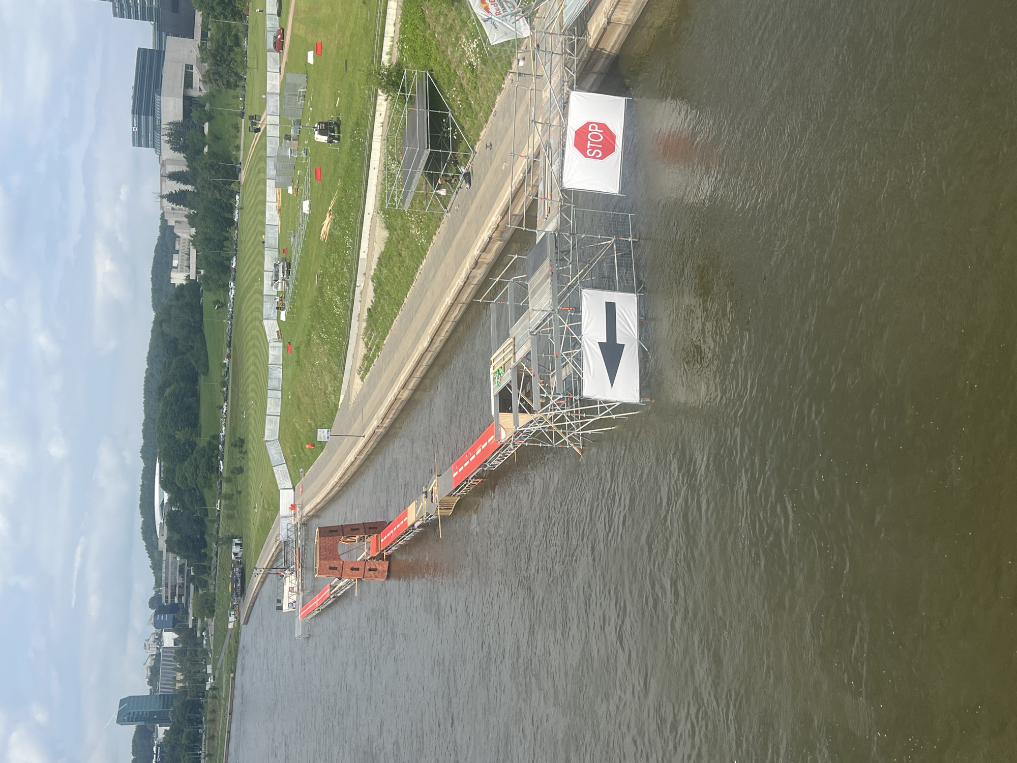 The image depicts a construction site on the Neris River in Vilnius, Lithuania. A temporary structure, appearing to be a bridge or walkway, is being assembled over the water. Red carpets are laid out on parts of the structure, and scaffolding surrounds it. A brick-like structure, possibly a decorative element or part of the bridge's design, is visible. Signs indicating a leftward arrow and "STOP" are hung from the scaffolding. In the background, a city skyline with modern buildings and green, tree-lined hills can be seen. The riverbanks are lined with paved walkways and grassy areas with some fencing and construction equipment present. The sky is overcast.