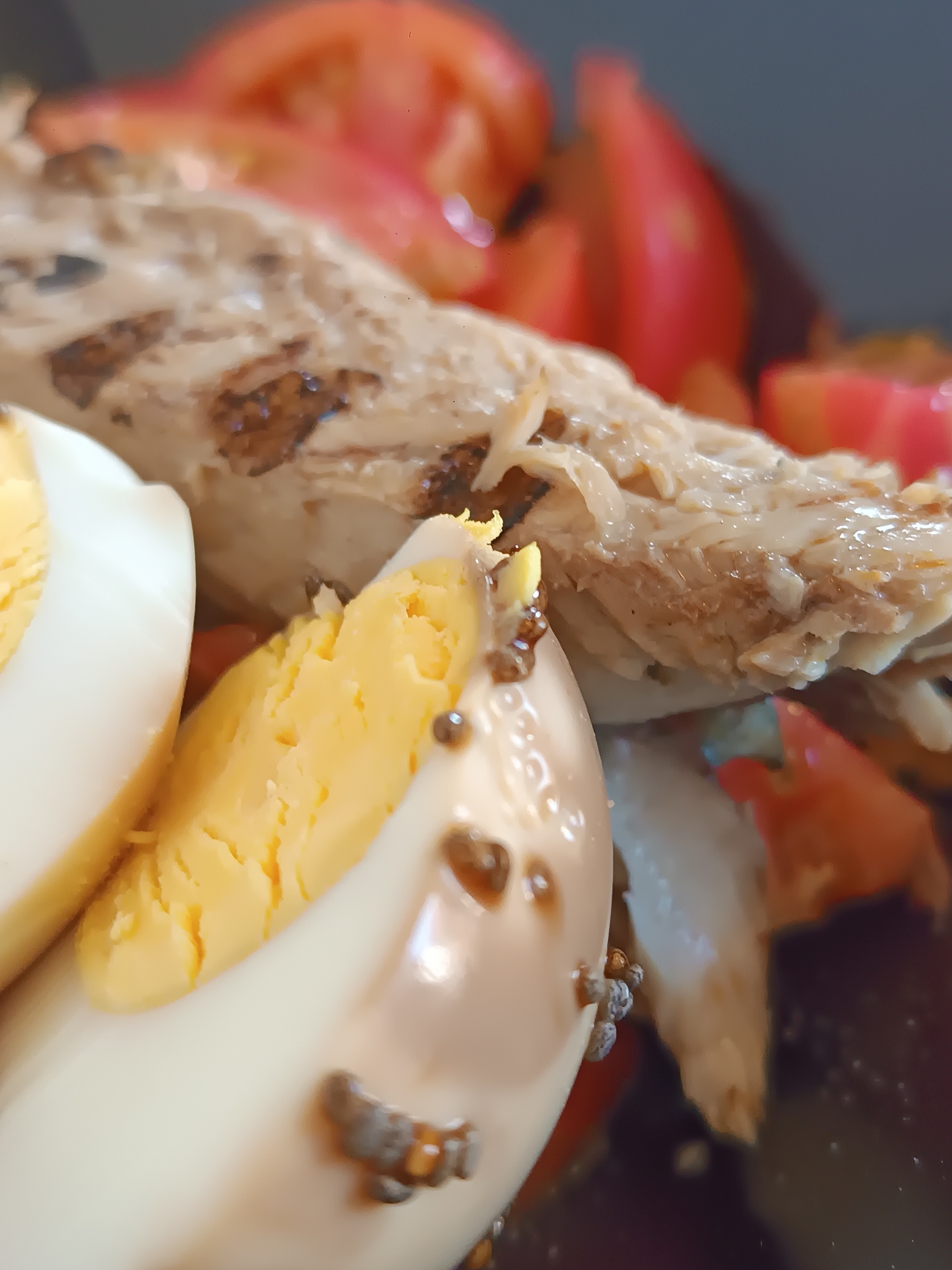 The image is a close-up, overhead shot of a plate of food. The foreground is dominated by two halves of a hard-boiled egg, with bright yellow yolks and white casings. Scattered on the egg white are small, dark specks, possibly pepper or herbs. Behind the eggs, a piece of grilled fish, flaked and slightly oily, is visible. The fish has grill marks and a light brown color. In the background, sliced red tomatoes are blurred, suggesting they are part of the same dish. The overall lighting is bright and direct, creating some glare on the food surfaces. The setting appears to be an indoor environment, likely a restaurant or home kitchen. There is no identifiable information regarding the location in Viadana, Italy.