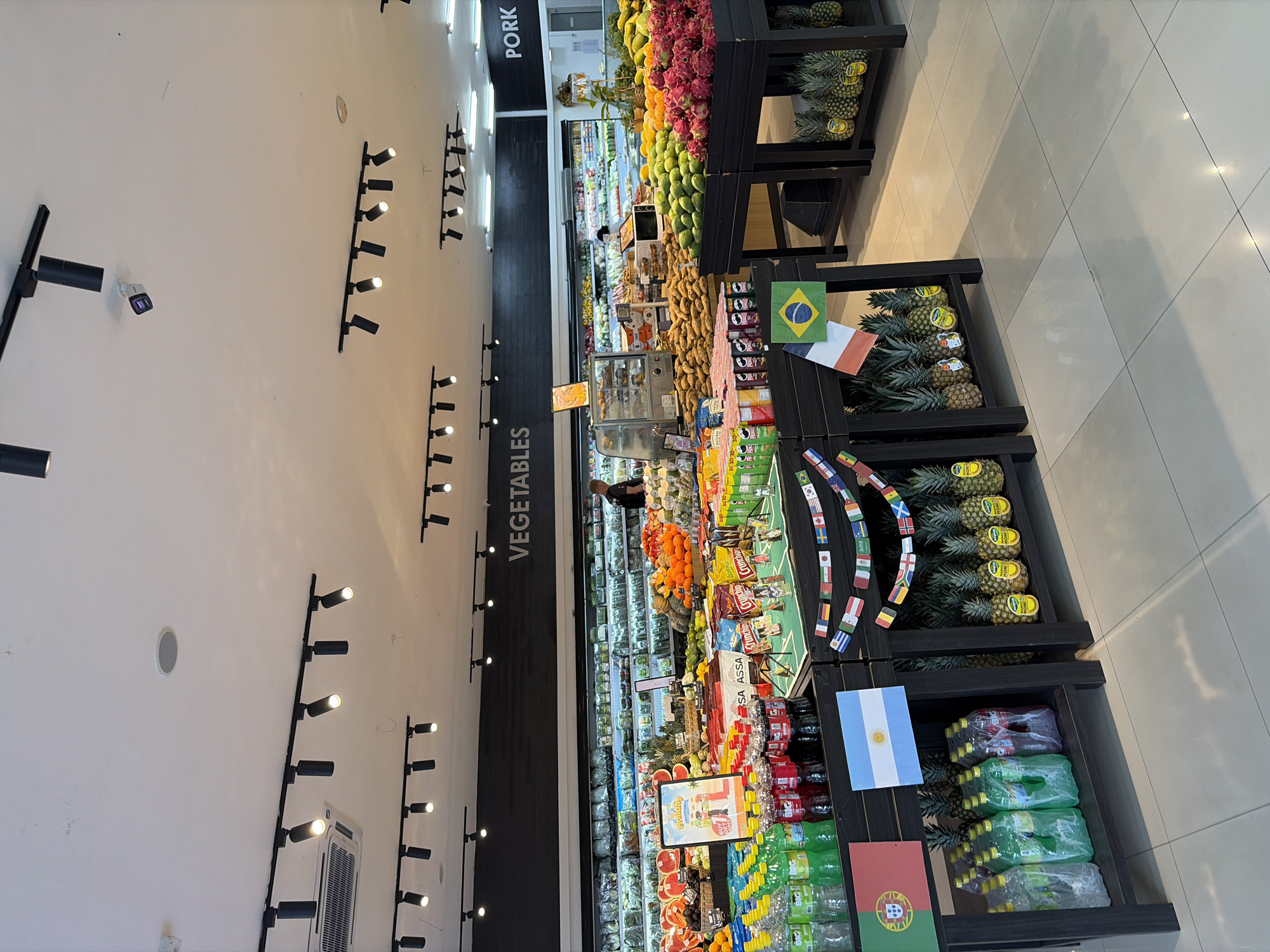 The image depicts the interior of a well-lit supermarket with a focus on produce and beverages. In the foreground, several black shelving units are arranged, showcasing a variety of items. On the left, a rack holds bottles of brightly colored beverages, some clear and some red. Above this rack, a flag with a shield emblem on a blue and white background is displayed, suggesting a possible theme or promotion. Further to the right, a string of small flags from various nations hangs across another shelf filled with pineapples.
Dominating the middle ground are displays of fresh produce. Piles of sweet potatoes, trays of dragon fruit, and arrangements of other fruits like mangoes and oranges are visible. Behind these produce displays, a long refrigerated section stocked with a wide array of vegetables is prominent. To the far right, a sign with the word "PORK" is visible on the wall, indicating another section of the store.
In the background, a store employee is visible near the refrigerated vegetable section, appearing to be engaged in stocking or organizing. The ceiling features numerous track lighting fixtures, illuminating the store. The floor is tiled with large, light-colored square tiles. The overall environment suggests a modern, organized retail space. The specific location in North Kuta, Indonesia, is inferred from external context not present in the image itself but provided as part of the prompt.