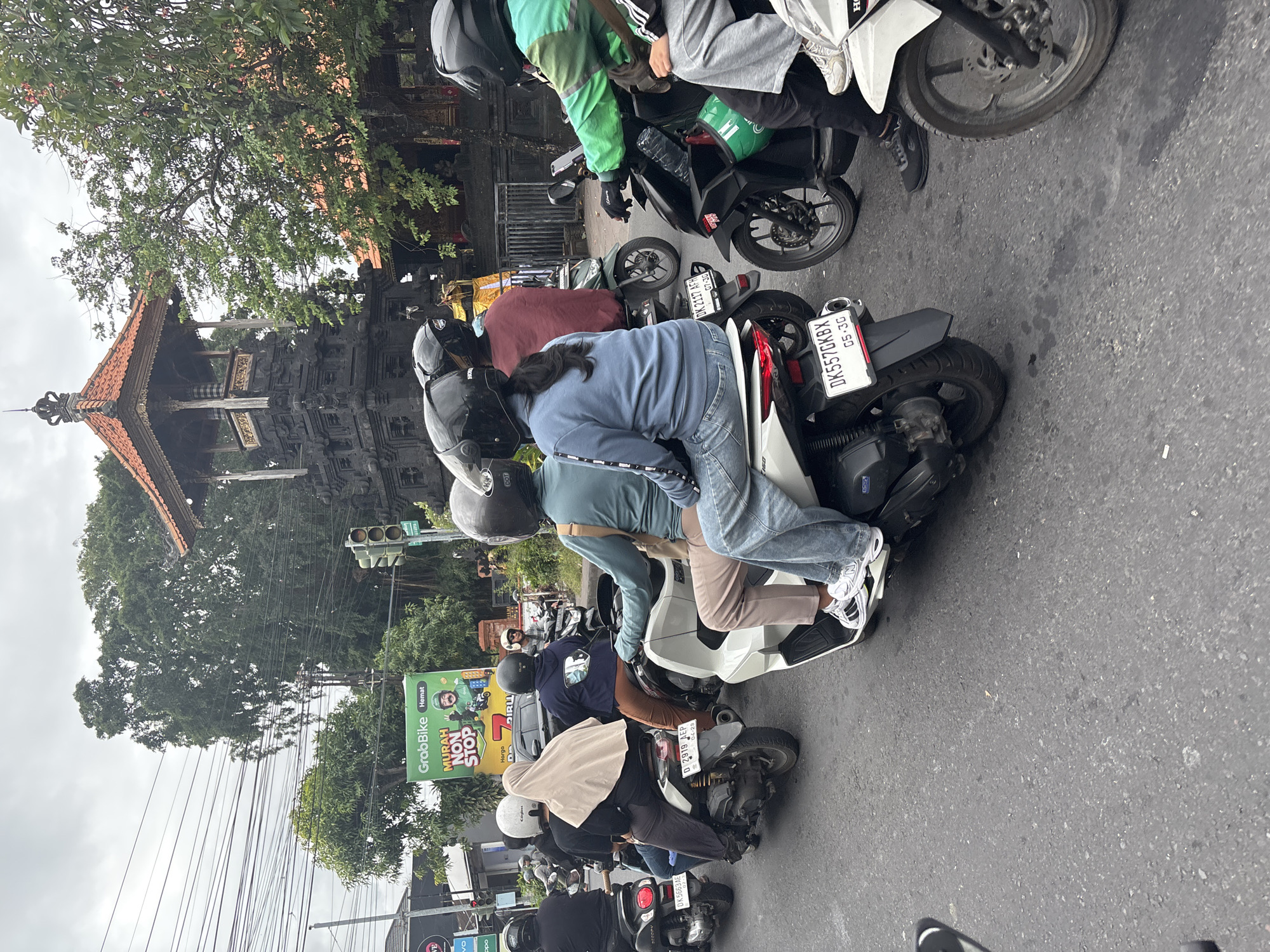 A medium shot captures a street scene in Denpasar, Indonesia, under an overcast sky, with a prominent religious structure in the background. Several people are riding on motorcycles, navigating a busy road. The foreground shows the rear of a white scooter with license plate "DK5570KBX". To its left is a black scooter with license plate "D 2919 AEP". Further left, a black scooter has license plate "DK 5663 AE". On the black scooter are two individuals, one wearing a dark shirt and the other a beige hijab. To the right of the white scooter, a black motorcycle has a green GrabBike bag and a water bottle on the handlebars. Another motorcycle to its right has a person in a green jacket. In the midground, a black motorcycle with two riders is visible, one with a dark shirt and the other with a red jacket. Behind them, a scooter with a rider in a blue hooded sweatshirt and jeans is also present. The background features a distinctive Balinese temple structure with intricate dark stonework and a tiered orange-tiled roof, surrounded by lush green trees. A large billboard for GrabBike, advertising "MURAH NON STOP", is also visible on the left side of the frame, near a traffic light. The road surface is asphalt, with some wet patches indicating recent rain. Overhead, a complex network of electrical wires crisscrosses the sky.
