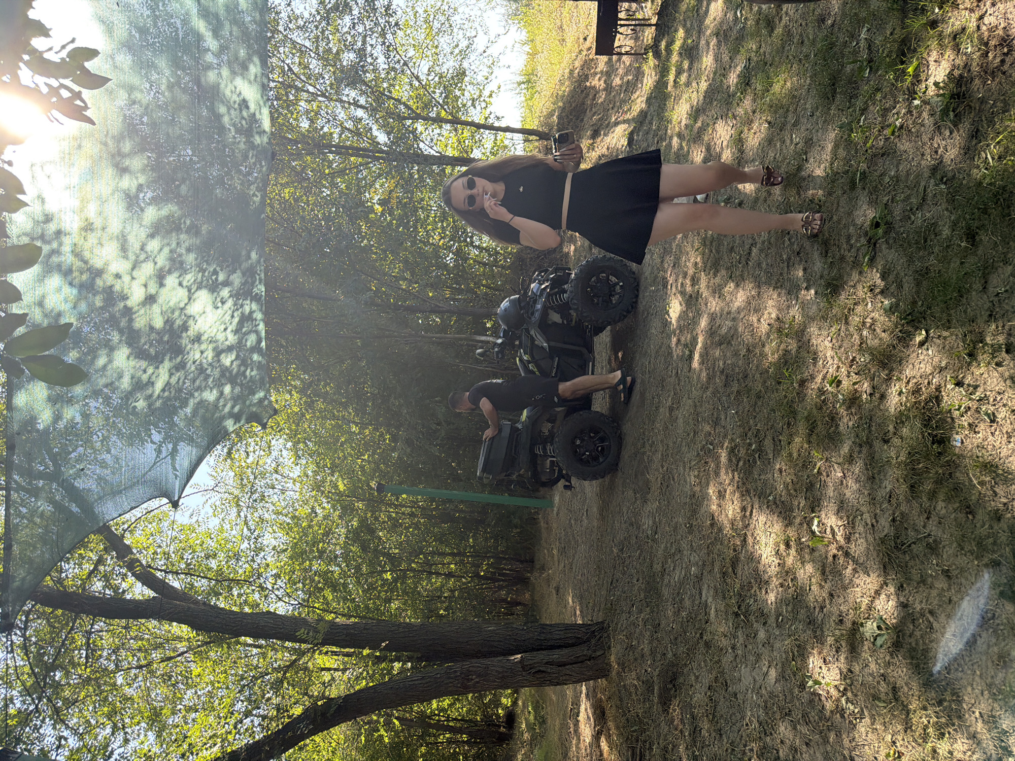 In Vatra, Moldova, on a sunny day, a woman in a black crop top and skirt walks on a dirt and grassy area, holding a small object and smoking. Behind her, an all-terrain vehicle (ATV) is parked, and a man in black shorts and a t-shirt is standing near it. A green mesh canopy is stretched overhead, casting dappled shadows. The background consists of trees and vegetation, with a bright sun visible through the leaves.