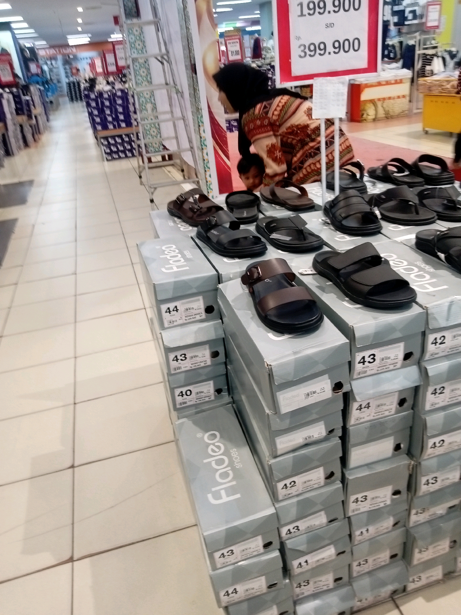 The image captures a retail store interior, likely a shoe section within a department store in Batam, Indonesia. The foreground is dominated by stacks of shoe boxes, some of which are open, displaying various styles of men's sandals. The boxes are uniformly grey with prominent size labels like "44," "43," and "40" printed in black. One open box in the foreground reveals a pair of brown sandals with a buckle closure. To the right, there's a stack of black sandals displayed on top of their boxes.
In the background, a woman wearing a dark hijab and a patterned dress is visible, bending over and appearing to interact with a child. A tall, silver ladder is positioned near the center of the image, leaning against a display. Further back, aisles of merchandise are visible, including what appear to be clothing racks and other retail displays. Signs with pricing information, such as "199.900" and "399.900," are displayed on a red and white signboard. The flooring is tiled and appears to be a light color. The overall lighting suggests an indoor retail environment.