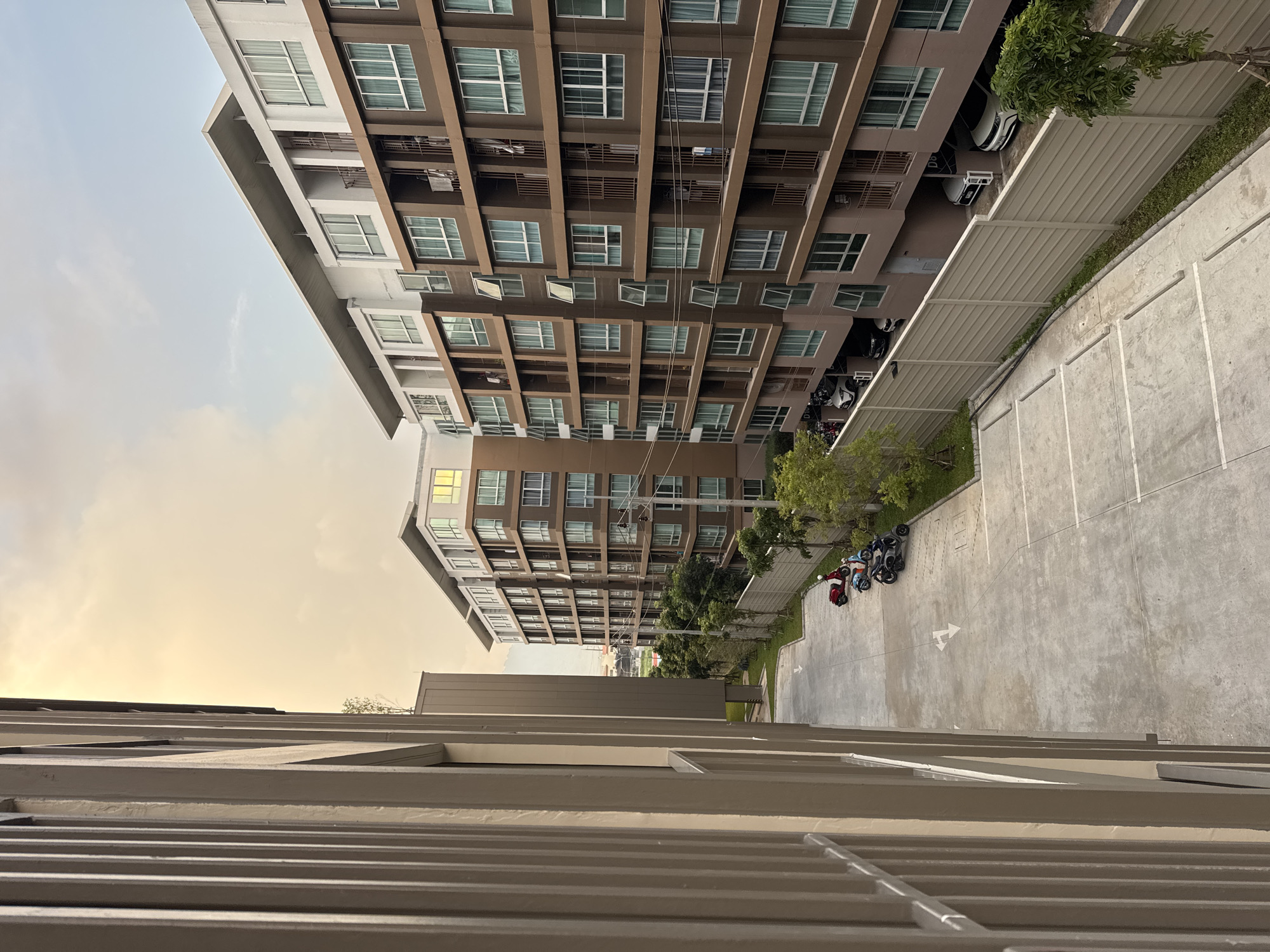 In Kathu, Thailand, an apartment complex with multiple levels features brown exteriors and white trim. The building's numerous windows are mostly closed, with some exceptions where they are open or their shutters are ajar. Balconies are visible on many floors. In front of the buildings, a paved parking lot is visible with several motorcycles parked in one section, and cars parked underneath the building in another. The parking lot is marked with white lines and directional arrows. A beige fence runs along the edge of the parking lot, separating it from a grassy area with trees. The sky above is a blend of soft clouds and warm hues, suggesting either dawn or dusk. The foreground is dominated by the side of another building with vertical, beige-colored siding.
