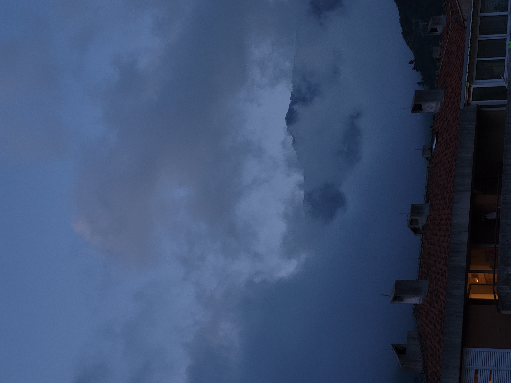 The image depicts a dusk or nighttime scene in or near Budva, Montenegro. A building with a red tile roof, windows, and ventilation structures is on the right side of the frame. One window is illuminated, suggesting the presence of indoor lighting. The background is dominated by a dramatic sky with heavy cloud cover, possibly indicating an impending storm. In the upper right corner, there are faint, dark hills covered in vegetation, which may be part of the mountainous terrain surrounding Budva. The scene suggests a residential area in Budva under a foreboding sky.