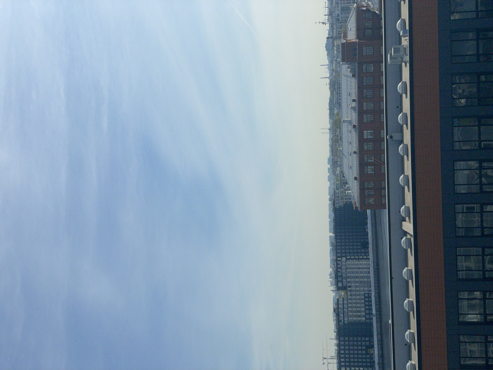 The image shows an urban landscape, likely a city, with a focus on buildings and the sky. The right side of the frame features the corner of a tall building with a dark blue facade punctuated by numerous rectangular windows arranged in a grid pattern. A strip of brown vertical paneling separates the blue facade from a light grey wall that is lined with dome-shaped protrusions, possibly skylights or architectural features.
Moving towards the center, a series of buildings are visible, decreasing in size and detail with distance, suggesting depth. The buildings vary in height, color, and design, ranging from dark, blocky structures to a lower, red-brick building with a glazed roof. The backdrop is a skyline with indistinct buildings in the far distance.
The left side of the image is dominated by the sky, which is a light blue with wispy white clouds. The sky gradually fades towards the horizon, becoming lighter and less distinct. The overall impression is one of a densely populated urban area with diverse architecture, viewed from an elevated position.