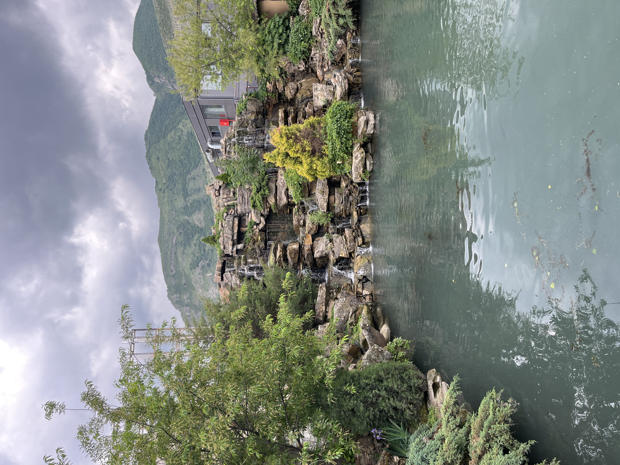 The image depicts a scenic view in Miatli, Russia. A rocky waterfall cascades down a landscaped structure adorned with various trees and bushes into a body of water. The water reflects the sky, which is overcast with dark, stormy clouds. A mountain looms in the background. A building is partially visible behind the waterfall, with a red flag-like object attached to it. In the foreground, there are trees and other greenery on the bank of the water.