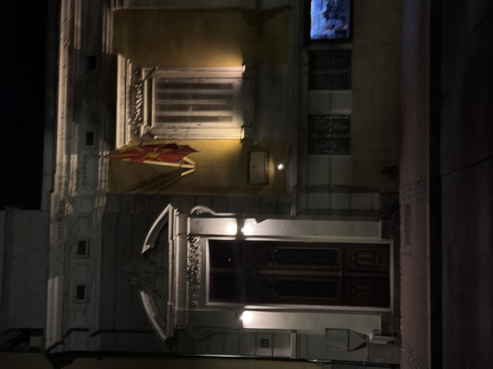 The image shows the facade of a building at night, likely an embassy or a governmental building, in Belgrade, Serbia. The building is dimly lit, with the exception of two bright lights illuminating the entrance. The entrance is a large, dark wooden double door, set within a white stone archway adorned with decorative carvings. To the left of the entrance, a house number "31" is visible. Above and to the right of the entrance, a window with closed shutters is lit from within by a warm yellow light, casting a glow on the facade. A flag, possibly the flag of North Macedonia, is hanging to the left of the window. Further to the right, a flat-screen television is mounted on the wall, displaying an image. The street in front of the building is paved with stone, and a dark road runs along the bottom of the image. The overall impression is one of quiet solemnity, with the illuminated elements highlighting the architectural features of the building.