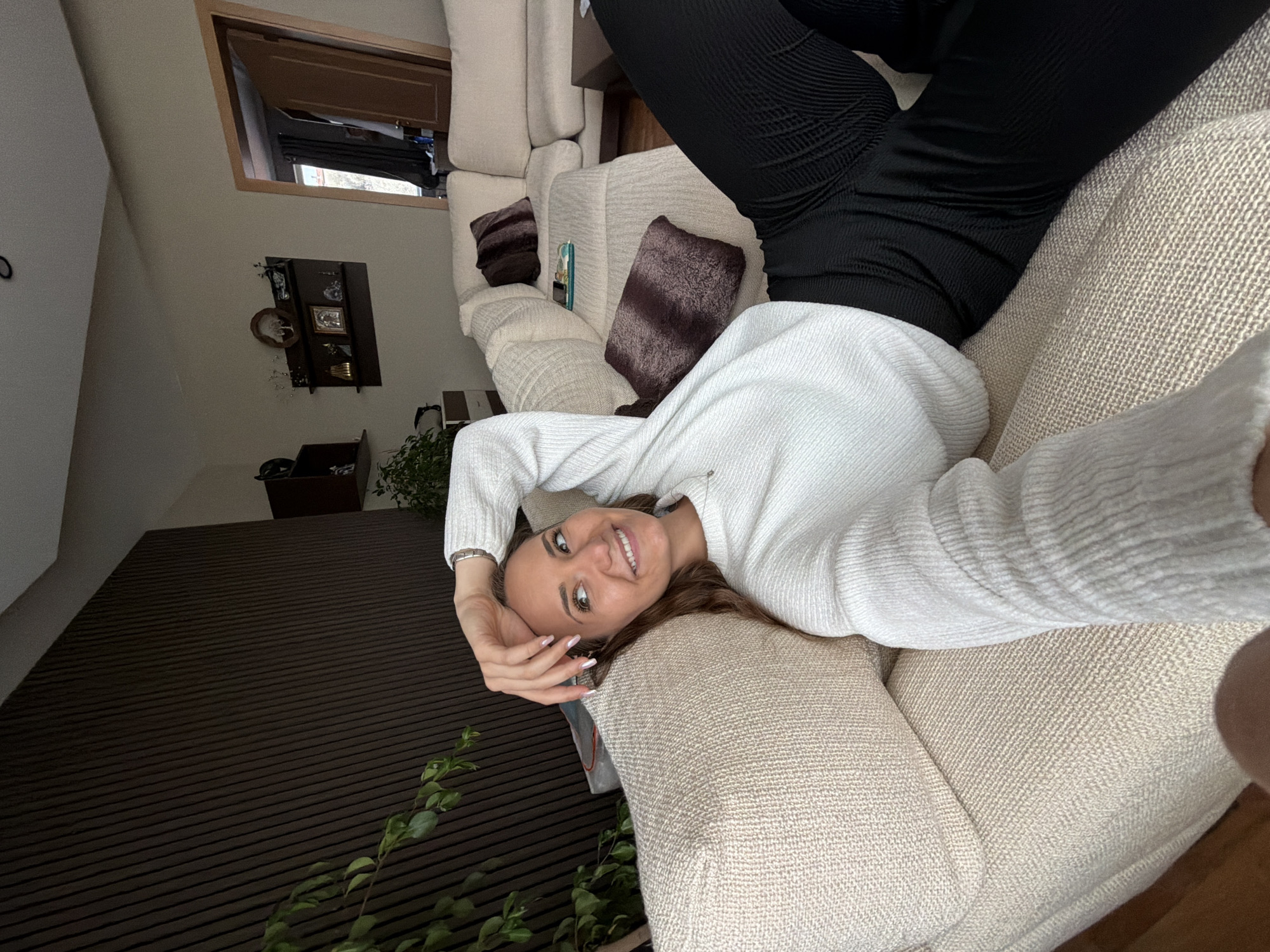 A woman wearing a white sweater and black pants is reclining on a beige sofa. She is smiling and looking at the camera. The background includes a dark brown wall with foliage, a window, and a shelf with decorative items. The location is unspecified but inferred to be a domestic interior. The city is not mentioned or visible in the image.