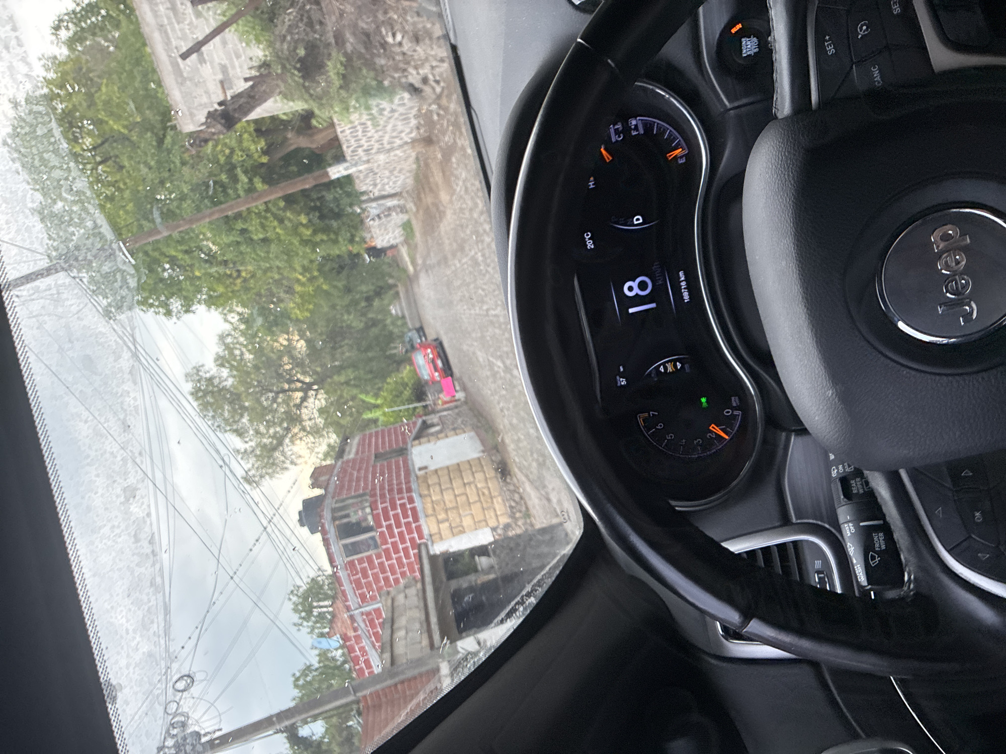The image, taken from inside a Jeep vehicle, shows a view of a street scene in Cañada de Madero, Mexico. The speedometer reads 18 km/h, and the temperature is 20°C. Outside the vehicle, there are buildings constructed with a mix of red brick and pale tan brick. Electrical wires run across the sky. The windshield shows signs of moisture or condensation. A red car is also visible on the street.