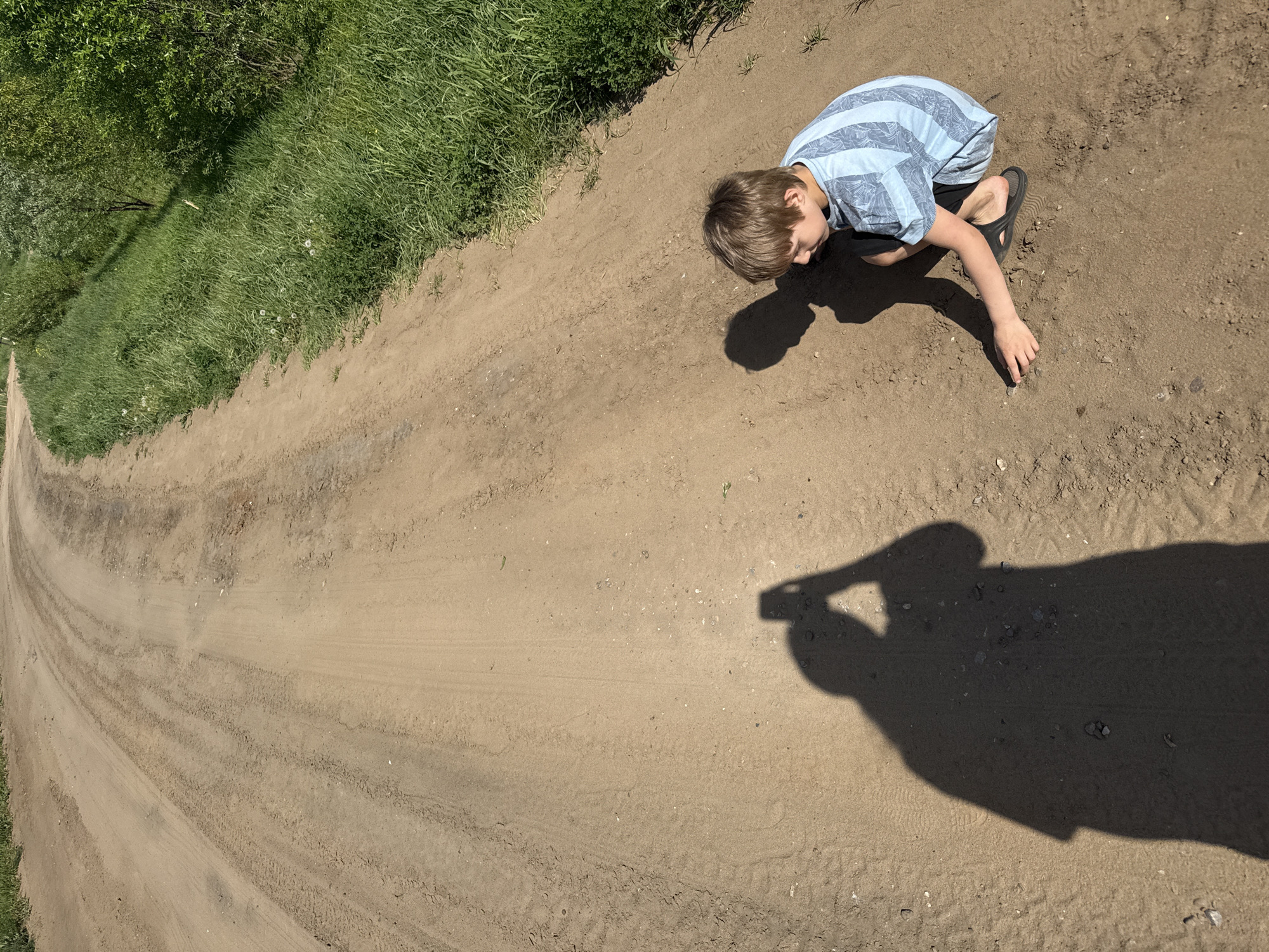 A boy wearing a blue and white striped shirt and black sandals crouches on a dirt road in Уральское, Russia. He is looking at something on the ground and reaching out to touch it. To the left of the boy, the dirt road curves into the distance, with grass and trees visible on either side. The shadow of someone holding a camera is cast on the road.