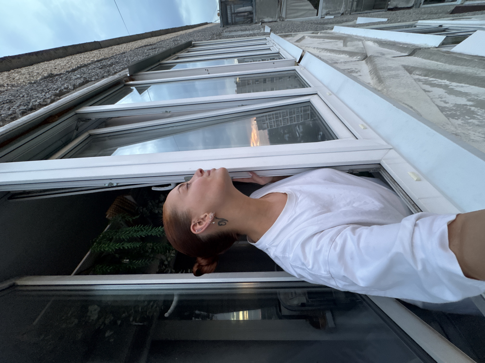 An individual with red hair pulled into a bun, wearing a white t-shirt, leans out of a window. They have multiple earrings in their left ear and a tattoo on their neck. The window overlooks an urban environment, with tall buildings visible in the distance and a cloudy sky. The building exterior appears to be made of rough concrete. The scene is likely a residential building balcony in Chaikovsky, Russia.