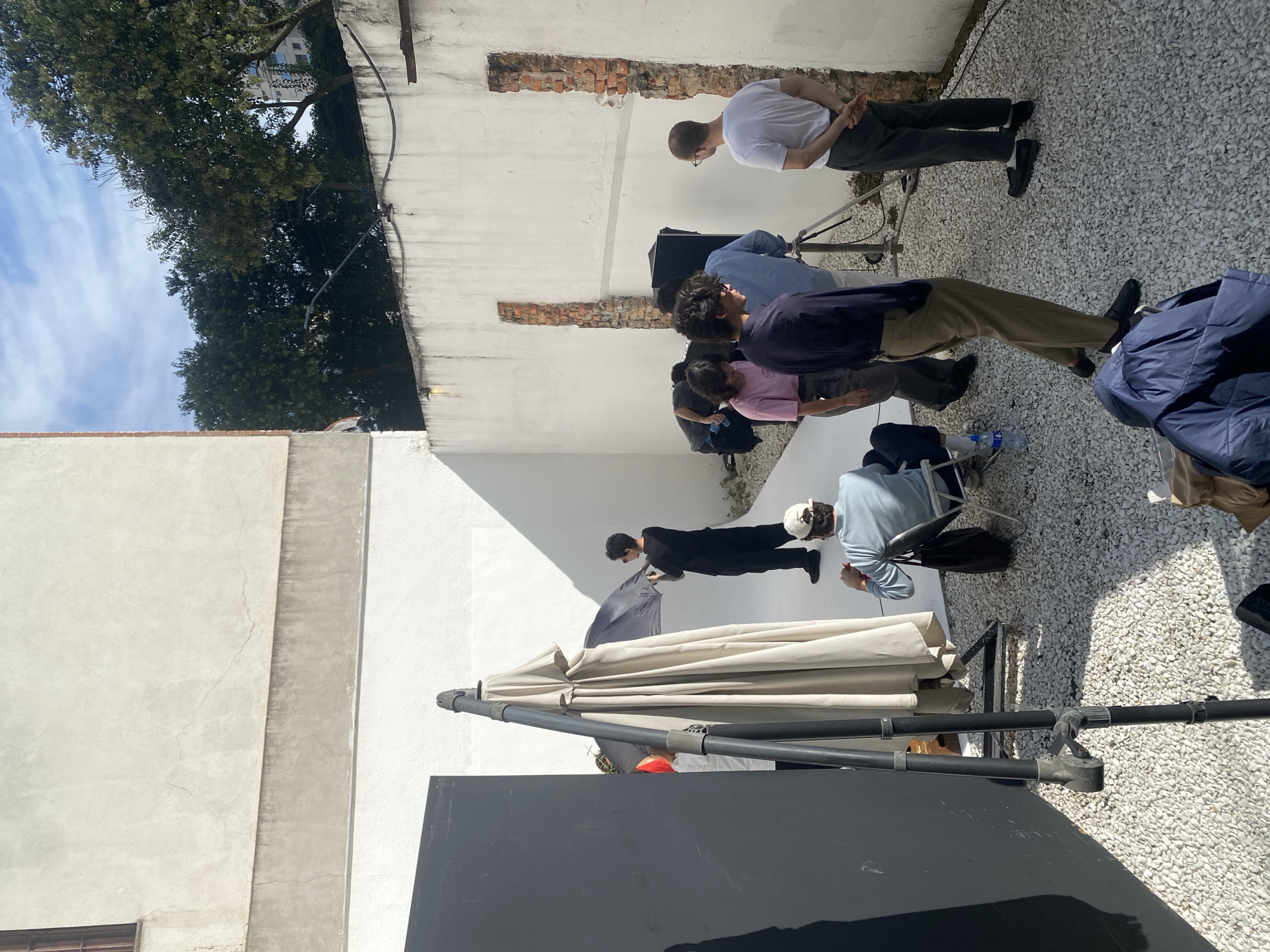 The scene appears to be a film or photo shoot taking place in an outdoor courtyard. The setting is a narrow area between two white-walled buildings with exposed brick sections. The ground is covered with white gravel. A group of approximately seven people are present, engaged in various activities related to the shoot. One individual in a white t-shirt and dark pants stands with their back to the camera, hands clasped behind their back. Another group of three individuals appears to be looking in the same direction, while two other people are sitting on chairs. One person is holding a reflector, and there is a large umbrella structure in the foreground partially obscuring the view. To the left, there is a building with a light gray facade. The sky is visible with some clouds. The location is the city of São Paulo, Brazil.