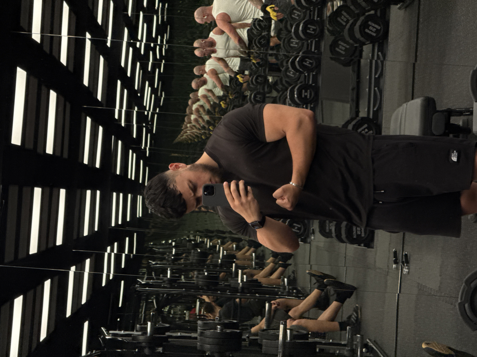 The image shows a man in a gym, taking a selfie in a mirror. He is wearing a black t-shirt and black shorts. The gym appears to be modern and well-equipped, with rows of dumbbells and weight machines visible in the background. The lighting is artificial, with long fluorescent lights running across the ceiling. The man is flexing his bicep for the camera. The reflection in the mirror shows the man and the gym environment. There is no discernible information in the image that can place the location in Varna, Bulgaria.