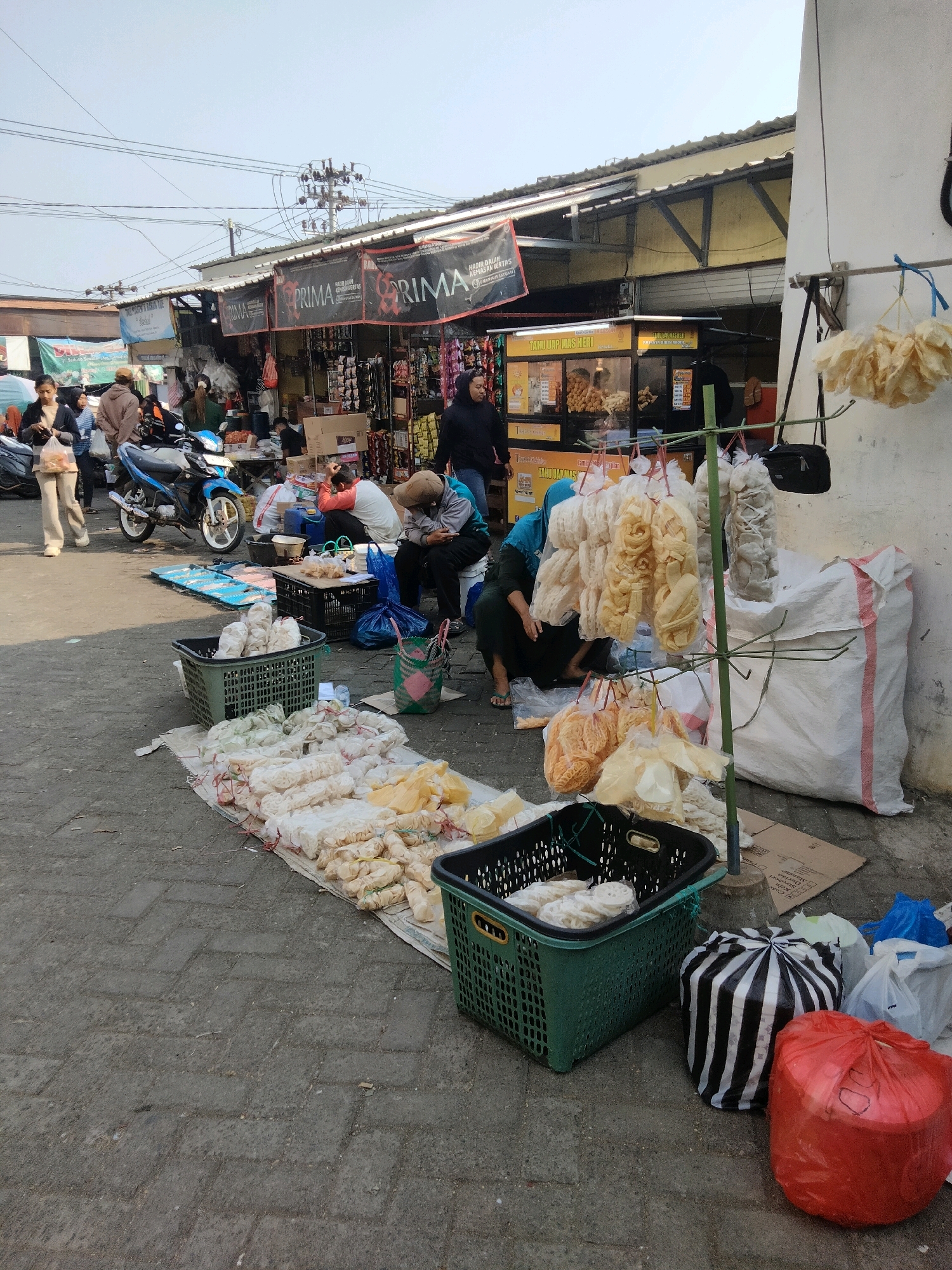The image depicts an outdoor market scene in Sawahan, Indonesia, with stalls lining a street. The foreground features a variety of packaged snacks, primarily crackers and puffed rice, displayed on mats and in green baskets. Some of these snacks are hanging on a metal rack. Several people are present in the image, some are customers browsing the stalls, and others appear to be vendors. In the background, motorcycles are parked, indicating this is a common mode of transportation. The stalls themselves are simple, with awnings providing shade, and appear to be selling a range of goods. The overall atmosphere suggests a typical day at a local market.
