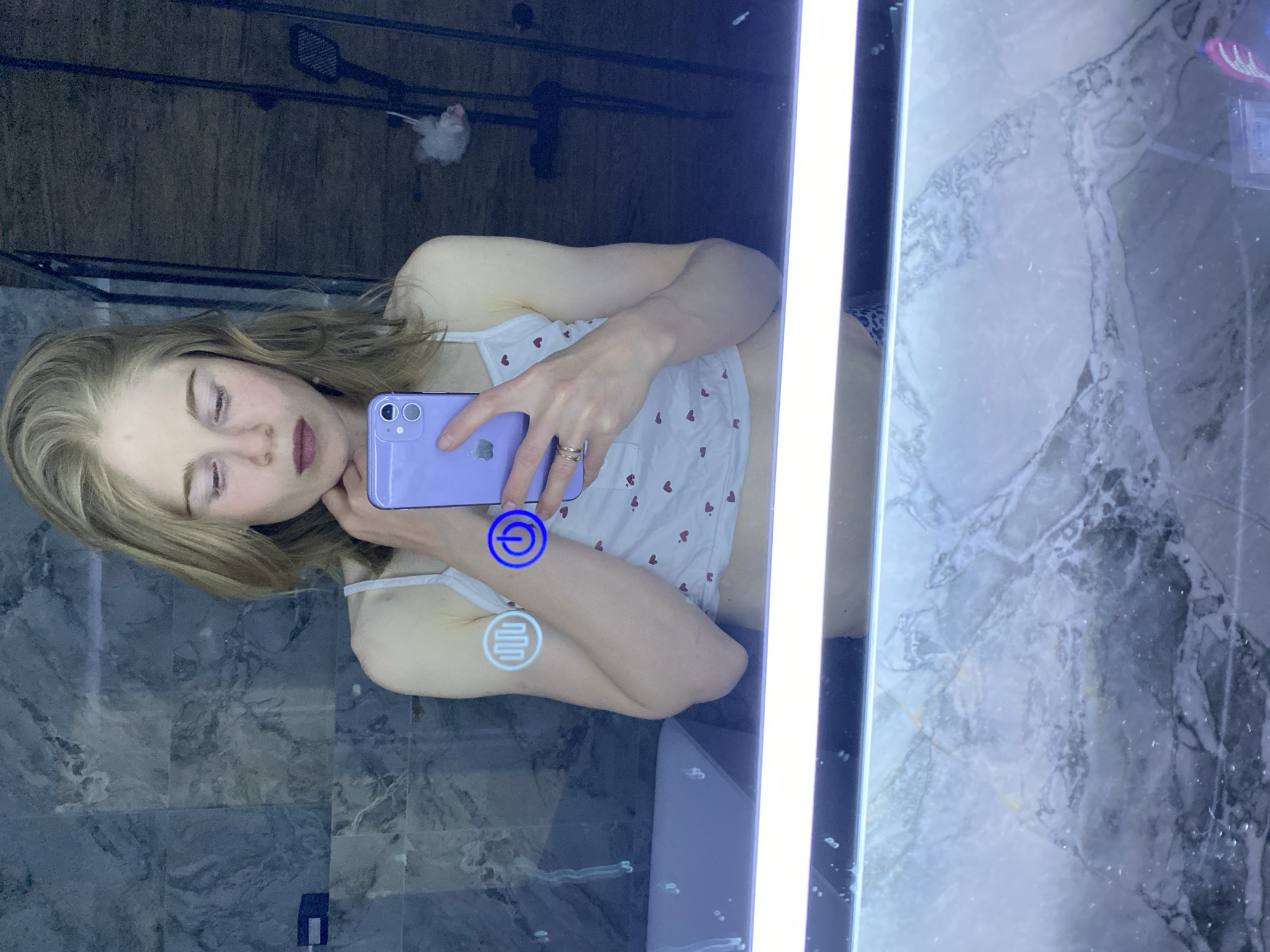 A young woman with blond hair is taking a selfie in a bathroom mirror. She is holding a purple smartphone. The mirror has a built-in light on the right side and touch-sensitive controls, a power button and a defogger. She is wearing a white tank top with red heart patterns. The bathroom has gray marble walls and a showerhead is visible in the background. The scene is potentially located in an apartment or home in Chaikovsky, Russia.