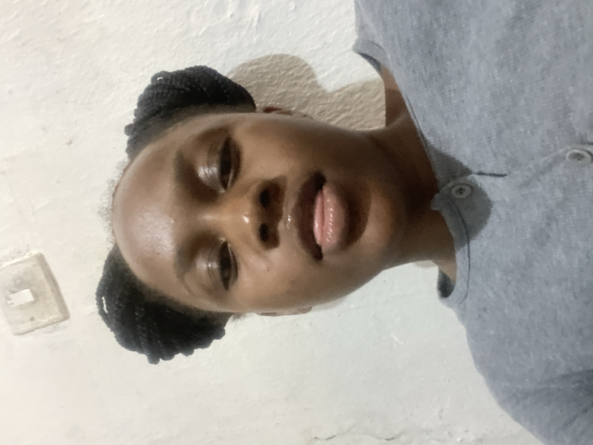 A young Black woman with dark braided hair in two buns is looking directly at the camera. Her face is illuminated from above, creating highlights on her forehead, nose, and cheeks. Her lips are parted, with her tongue slightly visible. She is wearing a gray crew-neck shirt. The background is a textured white wall. There is a light switch on the upper left side of the wall. The image appears to have been taken in an indoor setting. The location of Rafin Par, Nigeria is not visually ascertainable from the image.