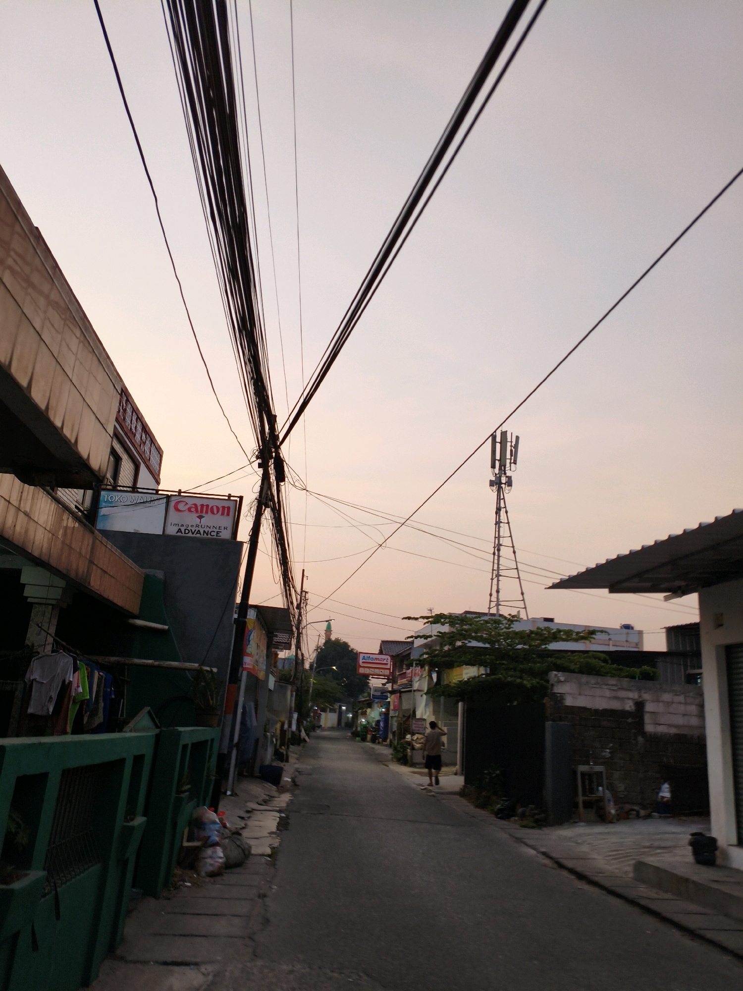 The image depicts a street scene in Bekasi, Indonesia, during what appears to be either sunrise or sunset, given the soft, warm light illuminating the sky. The perspective is from ground level, looking down a narrow street lined with buildings on both sides. Prominent overhead are numerous electrical wires, strung haphazardly and creating a dense network against the sky.
On the left side of the street, a two-story building is visible, with the lower floor featuring signage for "TOKO WAHYU" and "Canon imagerunner ADVANCE." Clothes are hanging on a clothesline in front of this building, suspended over a green metal fence. Adjacent to this, there is a pile of garbage bags.
The street itself is paved and appears to be moderately trafficked, with a single person walking away from the camera in the distance. Further down the street, a cell tower is visible. On the right side of the street, more buildings are present, some appearing to be shops with awnings. Another sign, "Alfamart," is partially visible. The overall impression is of a dense urban environment with a mix of residential and commercial structures, common in many Indonesian cities. The street is relatively clean, apart from the visible refuse on the left sidewalk.