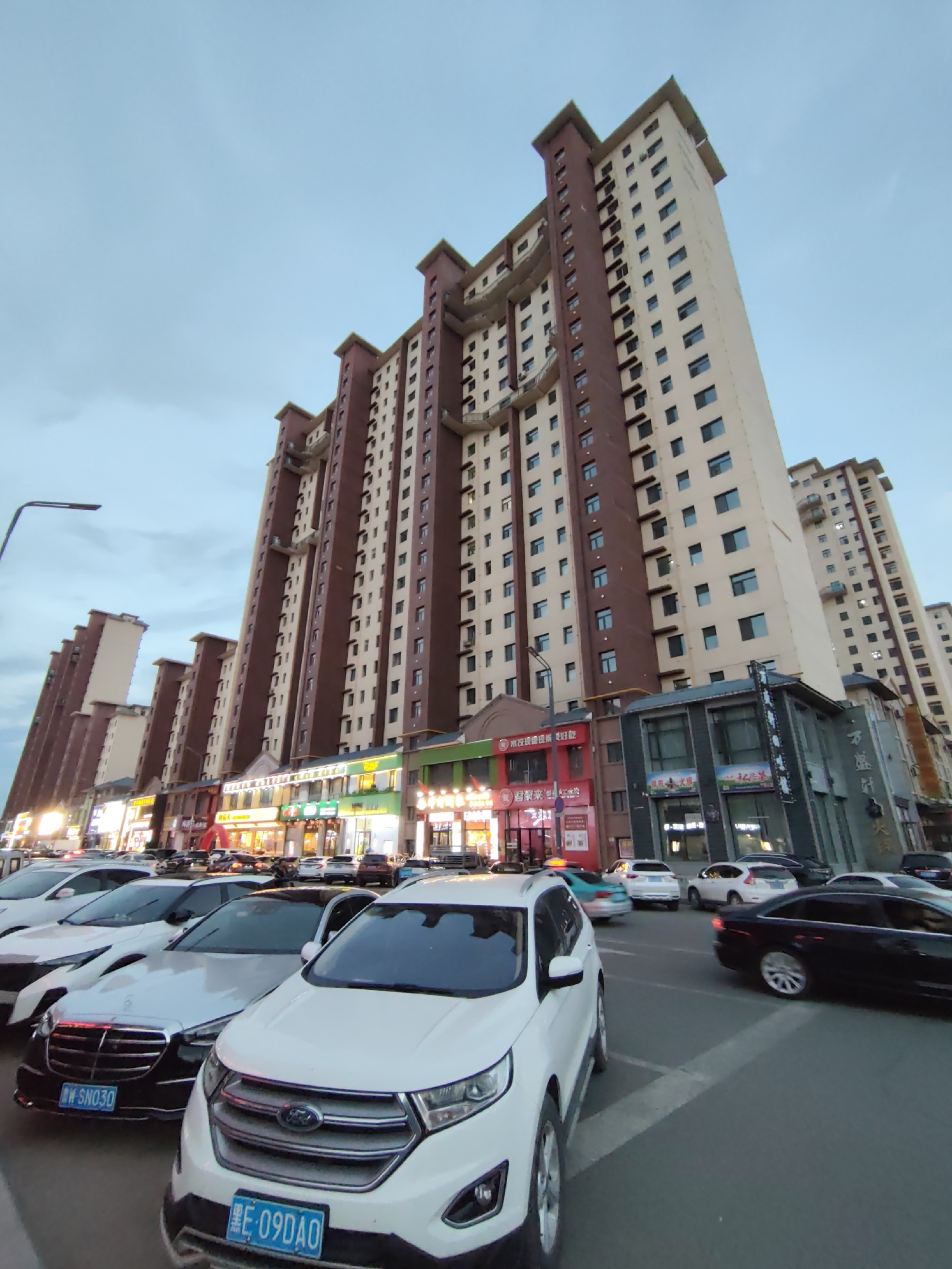 A low-angle, wide-shot captures a street scene in Lindian, China. The focal point is a row of tall, multi-story residential buildings with beige and dark red facades, stretching across the frame from left to right. The buildings are illuminated by the ambient light of the twilight sky and the glowing signs of the shops on the ground floor. Cars are parked and driving on the street in front of the buildings.
In the foreground, a white Ford SUV faces the camera. To its left is a dark-colored sedan with a license plate starting with "W SN030". Several other cars are visible in the middle ground and background, including a white Mercedes-Benz sedan, a silver sedan, and a teal-colored hatchback. The street appears to be a typical urban road with a white dividing line visible in the lower center of the frame.
The shops on the ground floor of the buildings feature bright, illuminated signs, suggesting a commercial area. Some signs have Chinese characters, indicating the location's origin. The sky is overcast with a pale blue hue, and a street lamp is visible on the left side of the image. The overall impression is a busy urban environment at dusk.