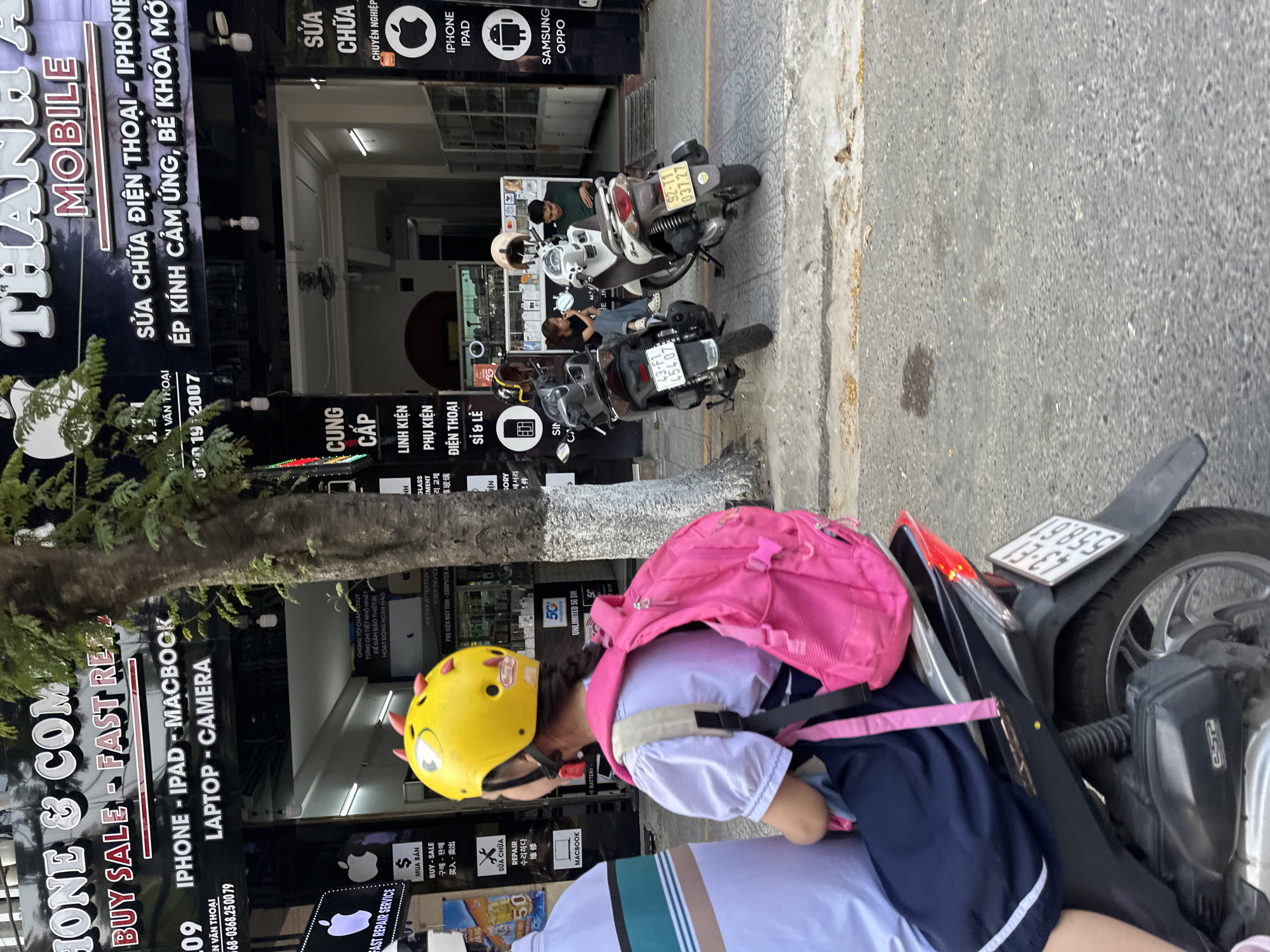 A person with a pink backpack and a yellow helmet is sitting on a black scooter parked on a sidewalk in front of several shops. The shops have signs advertising mobile phone repairs and sales, mentioning brands such as iPhone, iPad, and Samsung. Several other parked scooters are visible in the background. The scooter in the foreground has a license plate with the numbers 43-E3 198.59. The license plate on another scooter in the background is 43-F1 454.07. The location is in Đà Nẵng, Vietnam, as indicated by the license plate numbering system.