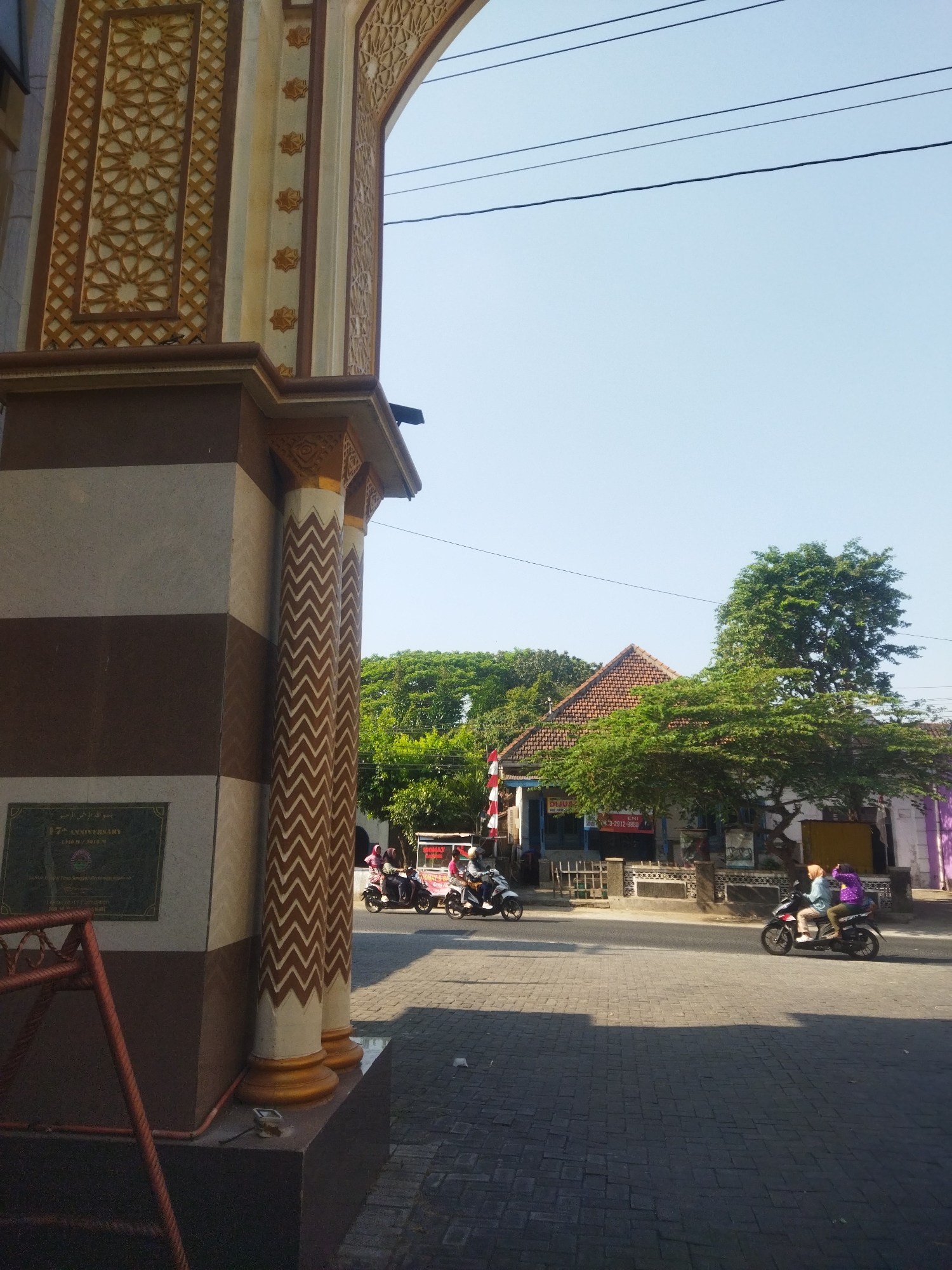 In Juwiring, Indonesia, an arched entryway with ornate Islamic geometric patterns on its facade frames a street scene. The entryway's pillars are decorated with a zigzag pattern in brown and white, and a plaque is mounted on the left pillar. Beyond the entryway, a two-story building with a red tiled roof is visible, bordered by trees. The street in front of the building is paved with dark cobblestones and asphalt. Several motorcycles are present: one is parked near the building, another is ridden by two people, and a third is ridden by a single person. The sky is clear and blue.