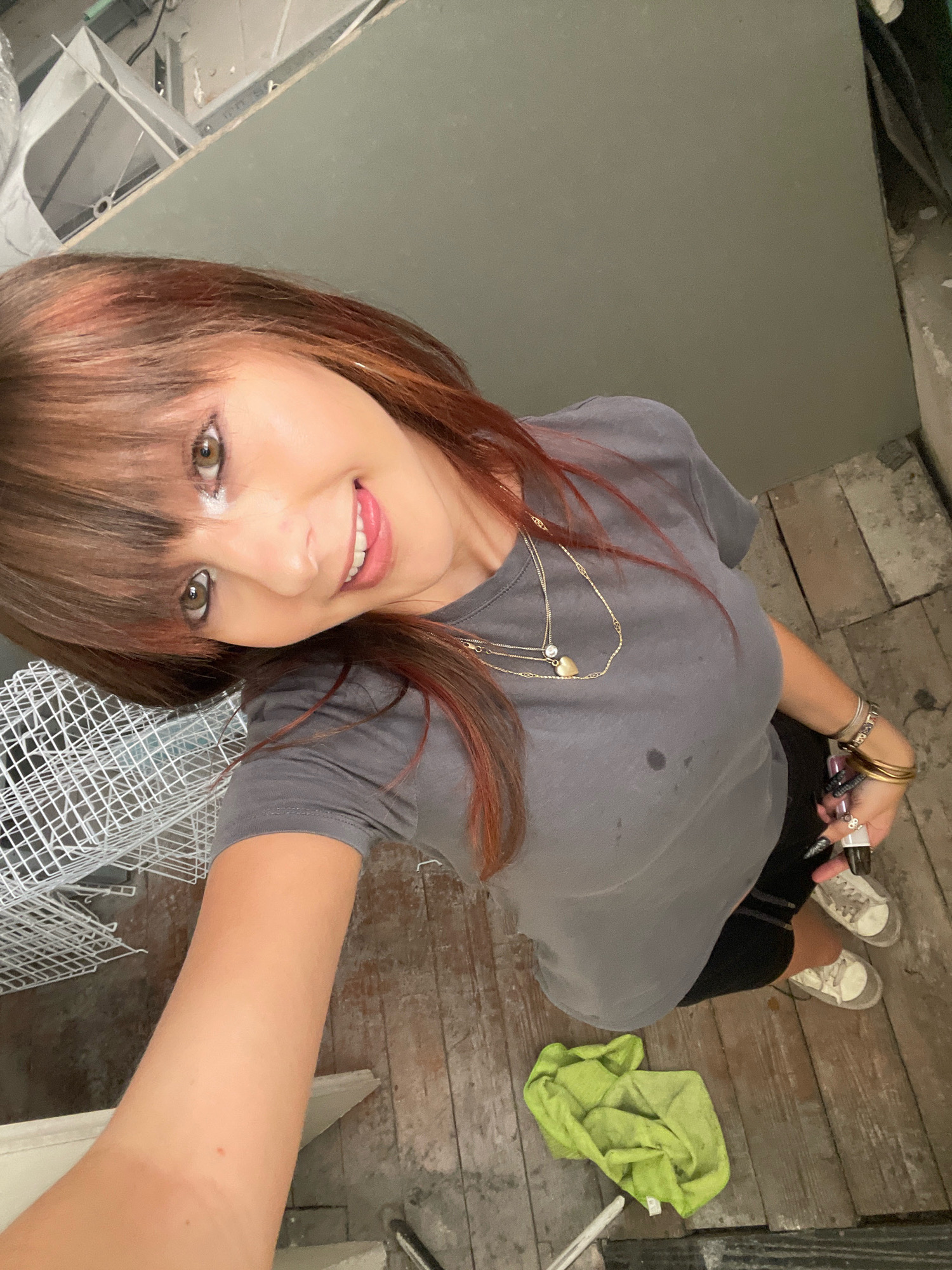The image captures a young woman from a high-angle, selfie-style perspective. She has auburn hair with bangs and is wearing a grey t-shirt, black shorts, and white sneakers. She is adorned with several necklaces and bracelets, and her fingernails are painted black and adorned with rings. The woman is smiling and her eyes are looking towards the camera.
The environment appears to be an indoor space undergoing renovation. The floor is made of weathered wooden planks, and there are exposed pipes and construction materials visible in the background, including what looks like scaffolding and drywall. A crumpled green cloth lies on the floor near her feet. The overall impression is one of an informal, perhaps candid, moment captured during a renovation project.
The location is identified as Raanana, Israel.