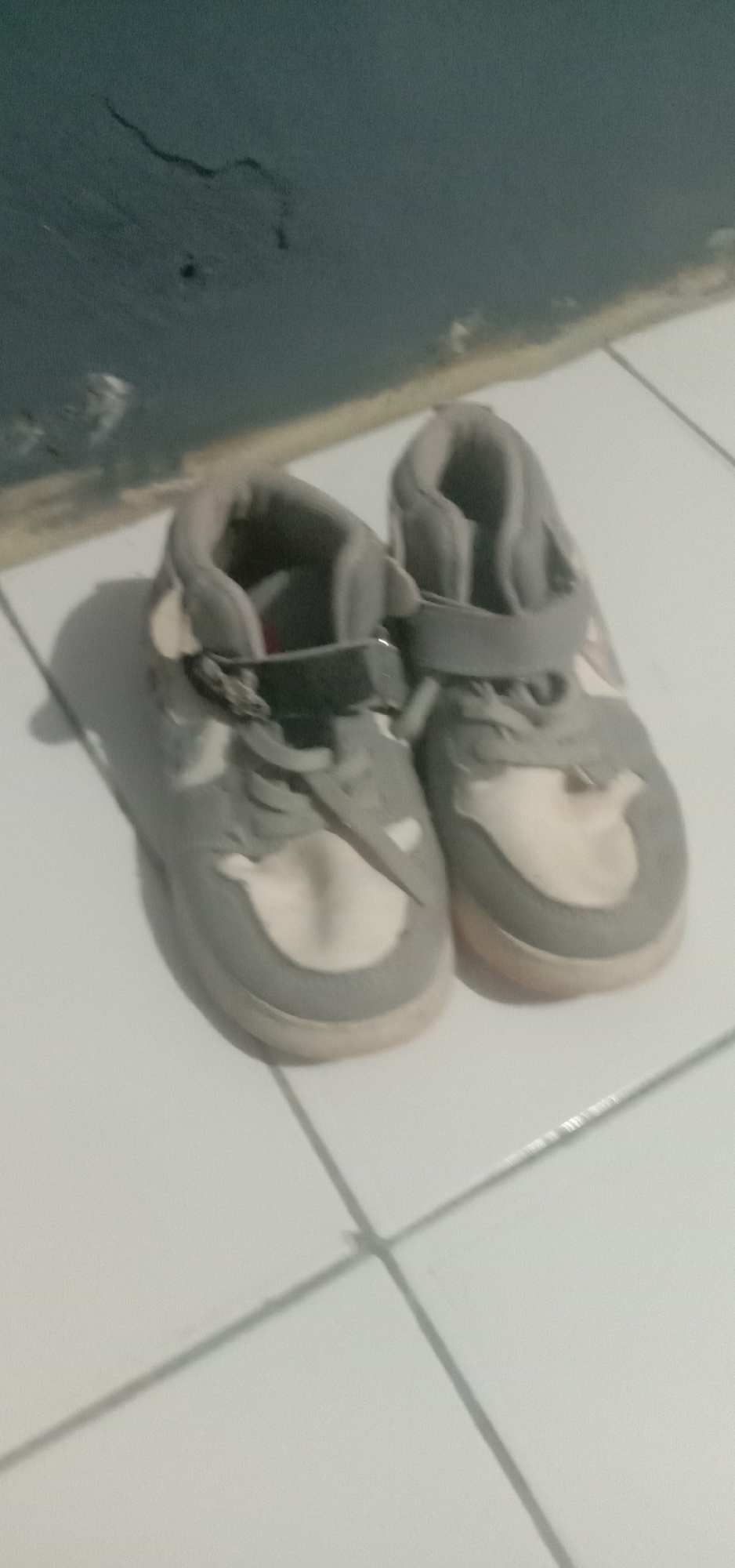 The image shows a pair of child-sized grey and cream sneakers placed on a white tiled floor. The sneakers have velcro straps and appear to be slightly worn. In the background, a dark-colored wall with some faint markings is visible. The lighting in the image is dim, casting soft shadows on the floor. The scene is indoors, likely in a residential setting. It is not possible to determine the specific location within Kendari, Indonesia, from this image alone, as there are no identifiable landmarks or contextual clues.