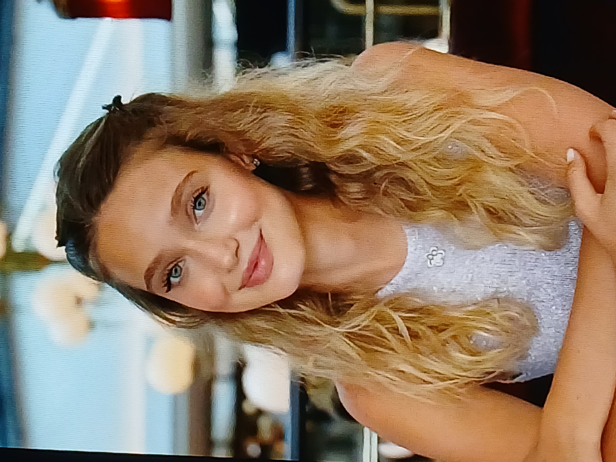 The image shows a close-up, waist-up shot of a young woman with long, wavy blonde hair and blue eyes. She is wearing a light blue, sequined top with a floral embellishment. Her head is tilted slightly, and she is smiling at the camera. The background is blurred, but appears to be an outdoor setting with soft lights. There is no visual information in the image that indicates the location is Dnipro, Ukraine.