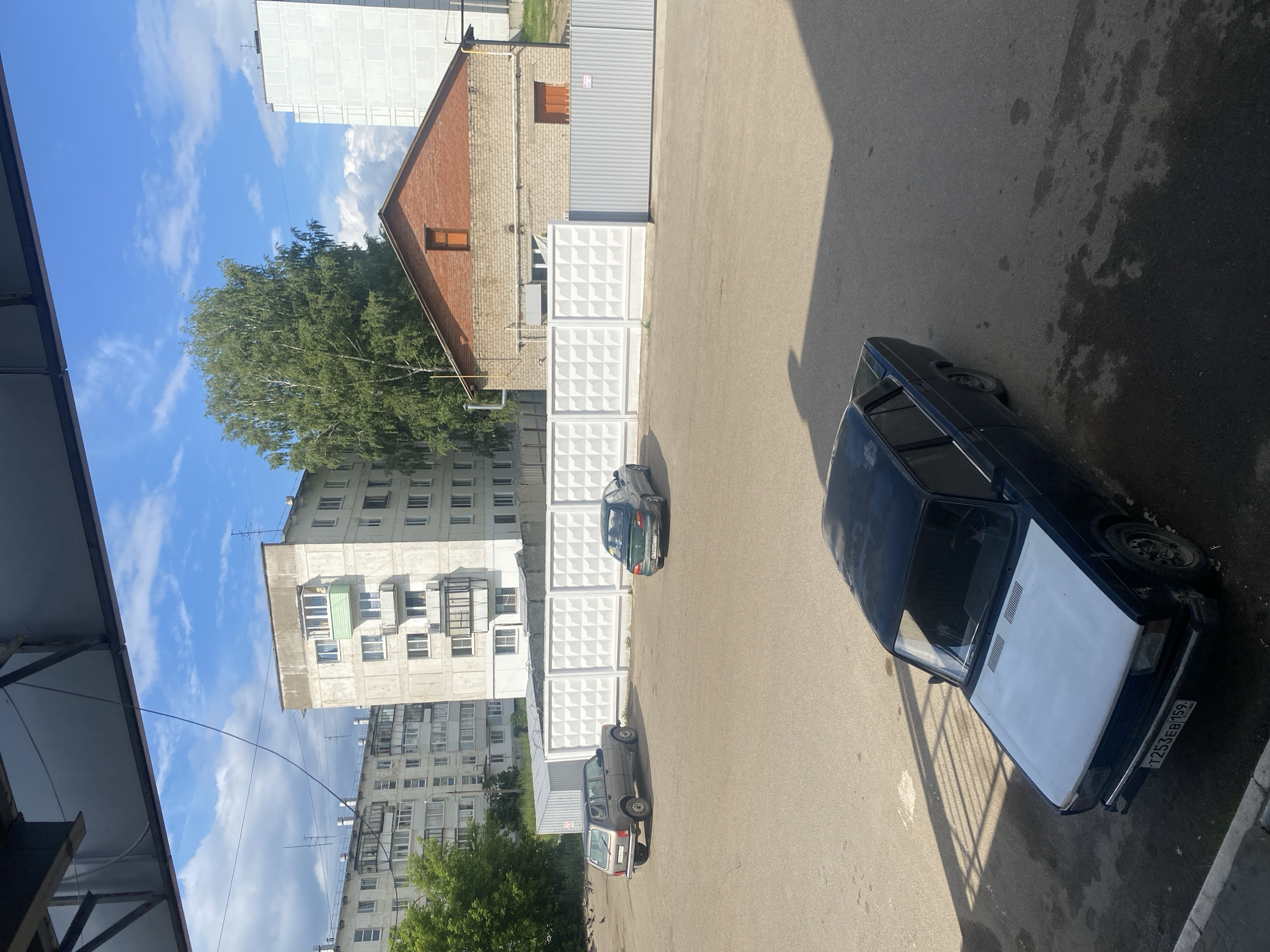 The image depicts a daytime outdoor scene in Chaikovsky, Russia, likely a residential area or an industrial courtyard. In the foreground, a dark-colored sedan with a white hood and a Russian license plate reading "T253EB 159" is parked on asphalt. In the middle ground, a gray SUV, possibly a Lada Niva, is parked to the left, facing away from the camera. To the right of the SUV, a dark green sedan is parked, also facing away. A white precast concrete panel fence separates the parking area from the background. Behind the fence, a red brick building with a brown wooden window is visible, along with a tall green tree. In the background, several multi-story apartment buildings, typical of Soviet-era architecture, are present under a blue sky with scattered white clouds. The overall environment appears to be an urban setting.