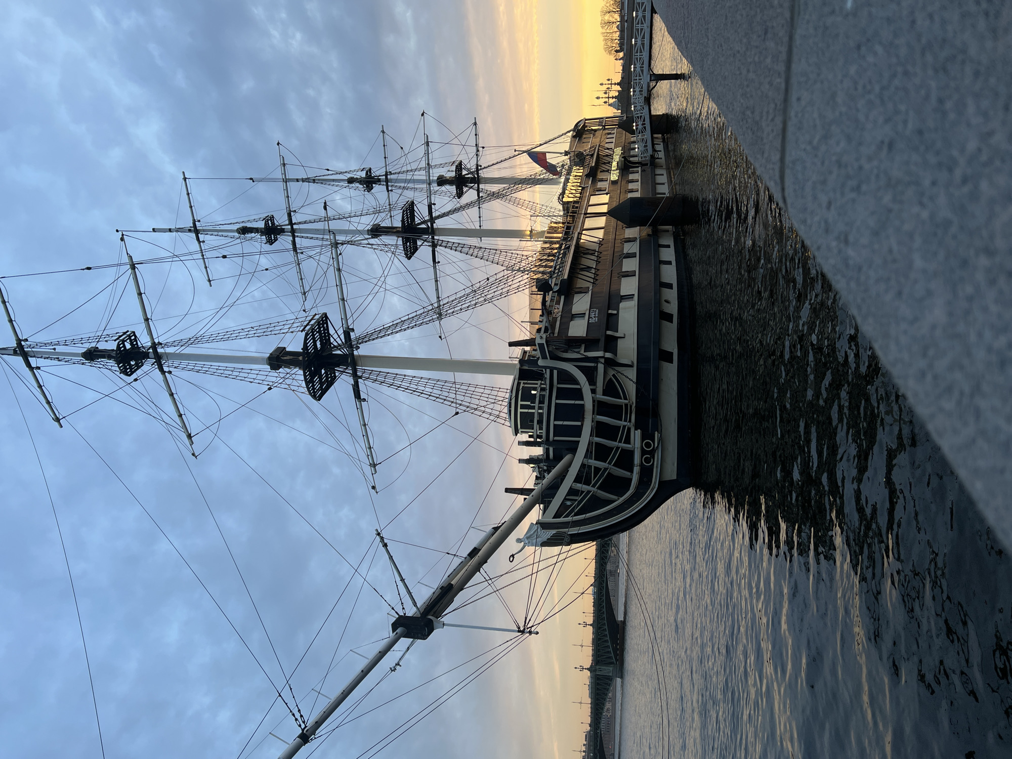 The image depicts a large, tall ship docked along a granite embankment in what is likely Saint Petersburg, Russia, given the architectural style and maritime history of the city. The ship, reminiscent of a historic sailing vessel, features tall masts and complex rigging. The hull is primarily black and white.
The ship is moored alongside a gray granite embankment that runs along the edge of the water. The water is relatively calm with visible ripples.
The sky is overcast with a hint of sunset colors near the horizon. A bridge can be seen in the background near the skyline.