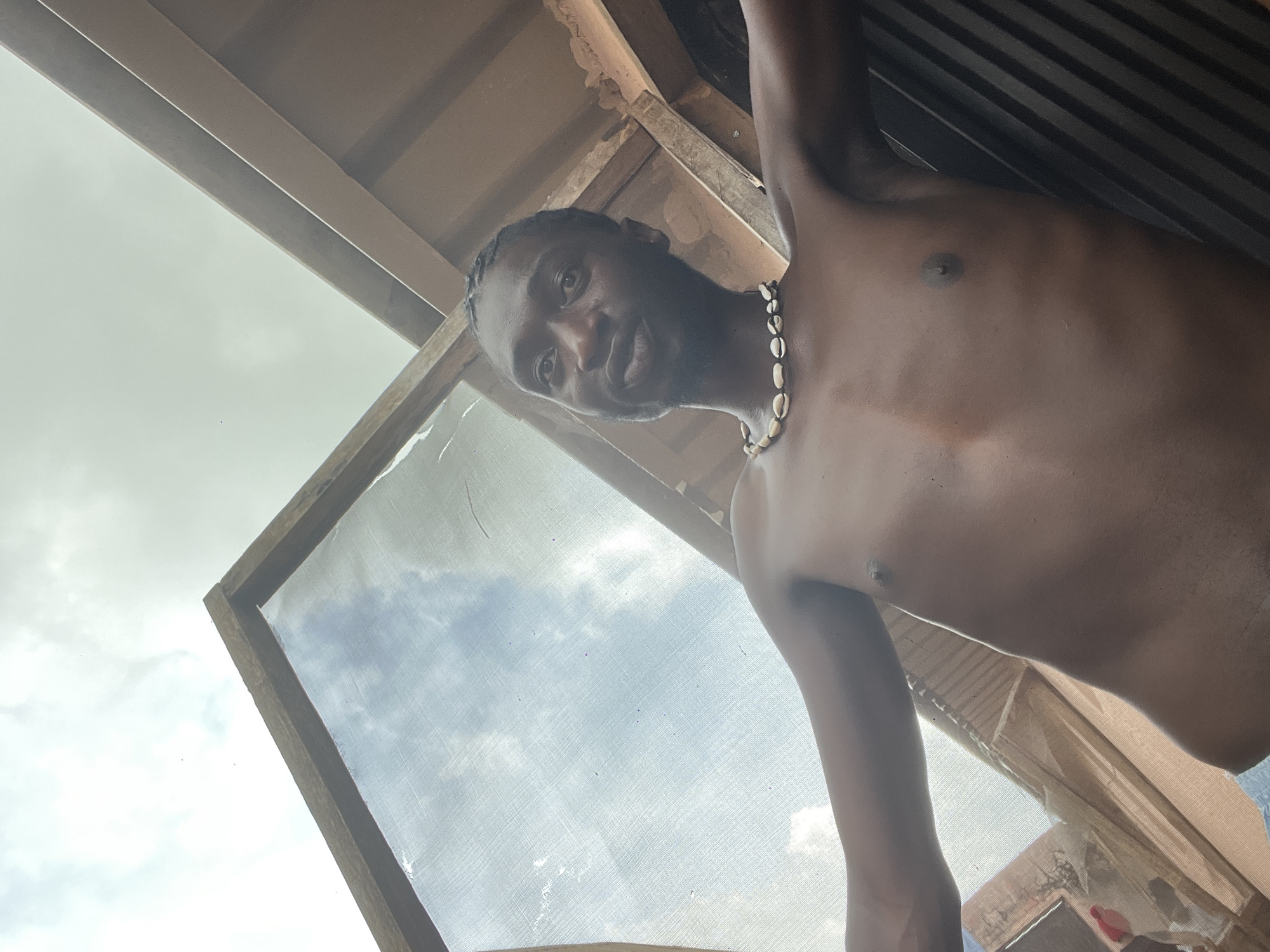 The image is a low-angle, close-up, outdoor shot of a shirtless Black man wearing a shell necklace. He is positioned in the foreground, facing the camera with a slight smile. The background shows a cloudy sky and some wooden structures that appear to be part of a roof or enclosure. The lighting suggests it is daytime. The image does not provide specific details about the location within the city of Durumi, Nigeria.