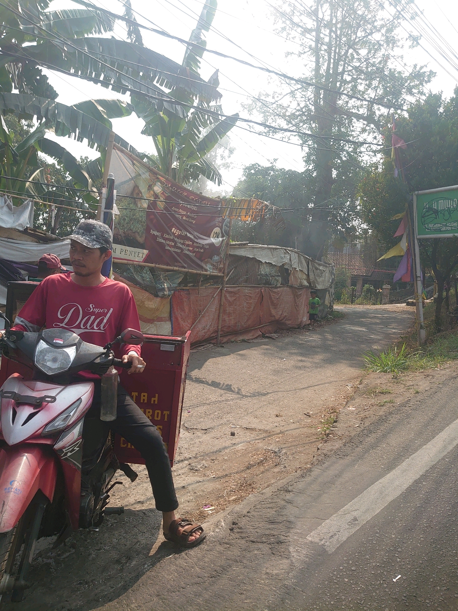 A man is sitting on a red motorcycle on a dirt road in Bojong Gede, Indonesia. The man is wearing a red t-shirt with "Super Dad" printed on it and a camouflage baseball cap. He is holding a cigarette in his right hand and has a bottle of water attached to the motorcycle's handlebars. The motorcycle has a large red storage container attached to the back. In the background, there are several stalls covered with tarps, some of which appear to be food vendors. A sign that reads "I U MUYU" is visible on the right side of the image. The scene is lit by bright sunlight, suggesting it is daytime.