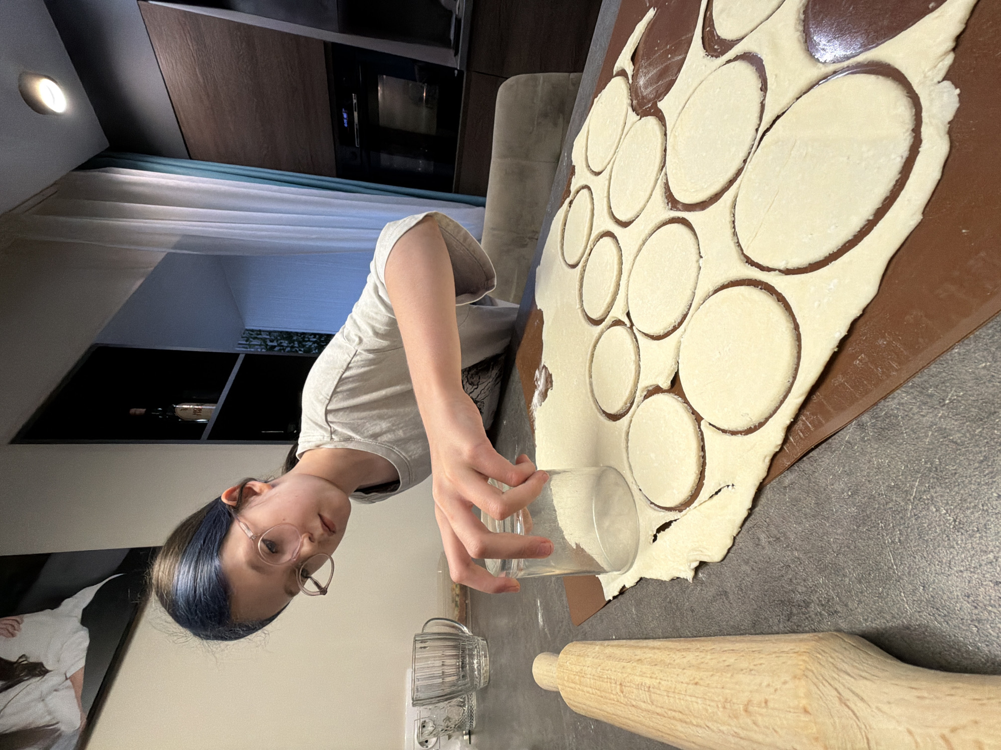 An image captures a young person engaged in baking or cooking preparation in what appears to be a kitchen or dining area in Birobidzhan, Russia. The shot is taken from a slightly elevated perspective and is rotated 90 degrees clockwise, showing the scene on its side.
The primary subject is a person, likely a young female, with light brown hair that has a noticeable blueish tint, wearing glasses with round, light-colored frames. She is dressed in a simple, light-colored t-shirt. Her right hand is holding a clear glass, which she is using to cut circular shapes from a sheet of rolled-out dough. The dough rests on a brown silicone baking mat or similar surface, with several circular pieces already cut and the excess dough remaining around them.
To the right of the dough, a wooden rolling pin is visible on a dark grey countertop, suggesting it was recently used. In the foreground, below the person's hand, a clear, ridged glass mug sits next to a white power outlet on the same countertop.
The background reveals a modern interior. Behind the person, a dark wooden cabinet and a built-in oven with a dark reflective surface are visible. A light-colored, comfortable-looking armchair is partially seen. Further back, light-colored curtains with a teal accent strip are drawn, indicating an indoor setting. A wall-mounted light fixture casts a warm glow, suggesting it might be evening or night. On a shelf or wall behind the person's head, what appears to be a framed picture or artwork is partially visible. The person appears focused on the task, with no strong emotions discernible. No visible text is present in the image.