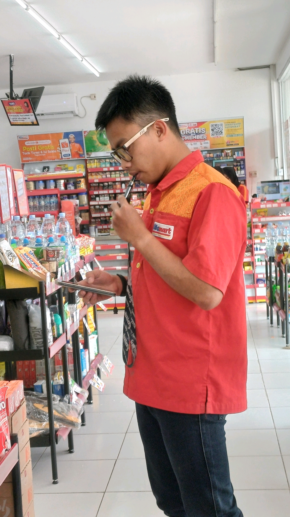 A man wearing a red Alfamart uniform is standing in an aisle of a convenience store, looking down at his phone while holding a vape pen to his mouth. The store shelves are stocked with various products, including bottled water, snacks, and personal care items. The man's uniform has a prominent Alfamart logo on the chest. The environment appears to be a typical Indonesian convenience store, with fluorescent lighting and tiled floors.