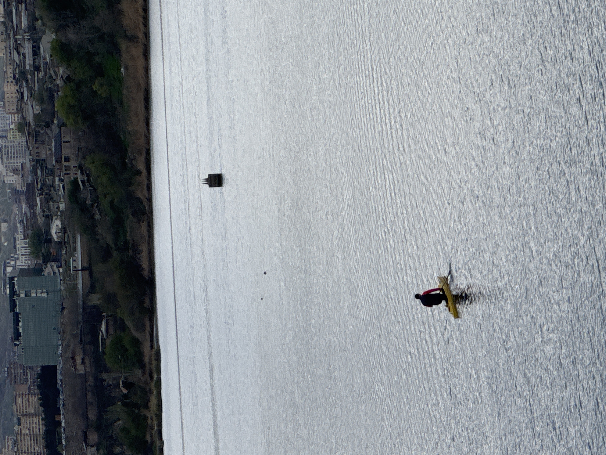 The image presents a high-angle view over what appears to be a large body of water in Yerevan, Armenia. Along the left side of the water are buildings and vegetation suggesting a city edge. In the water, there's a small rectangular structure close to the left bank. Further out in the water, a person is on a yellow surfboard. The water surface shows subtle ripples, indicating some wind activity.