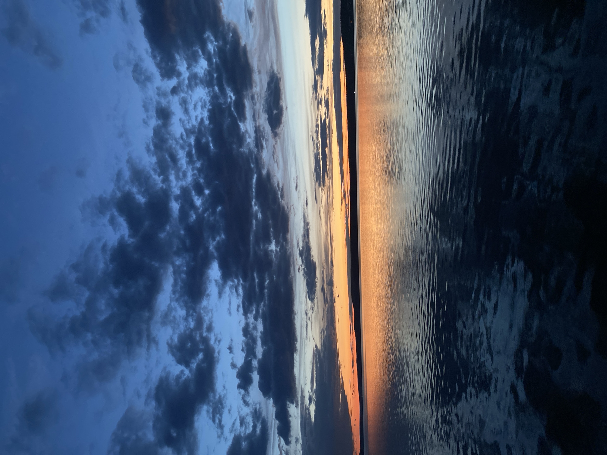 The image depicts a sunset view over water, possibly a lake or river. The sky is filled with dark, textured clouds, transitioning from a dark blue at the top to lighter shades near the horizon where the sun is setting. A band of orange and yellow light marks the location of the setting sun. The water surface reflects the colors of the sky, with a darker, more muted palette towards the bottom of the frame. The water shows small ripples and reflections of the sky.
Without additional context, it is impossible to confirm the exact location within Votkinsk, Russia. However, based on the presence of water and sunset, it is possibly on the shore of the Votkinsk Reservoir or a similar body of water within or near the city.