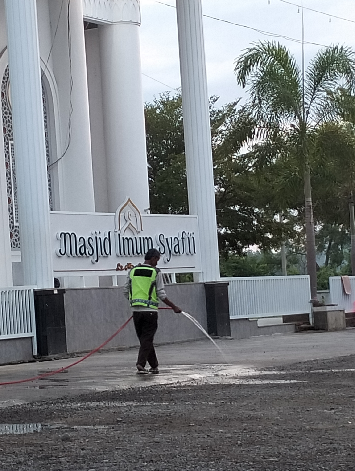 In Blangmetimu, Indonesia, a person wearing a bright yellow-green safety vest and dark pants is seen from behind, walking on a wet, gravelly surface. The individual is holding a red hose from which a stream of water is spraying onto the ground. To the left of the scene, a large white building with columns and a sign that reads "Masjid Imum Syafii" is visible. The environment appears to be an outdoor area adjacent to the mosque, with palm trees and other foliage in the background under an overcast sky. The ground is a mixture of asphalt and gravel, with puddles reflecting the sky.