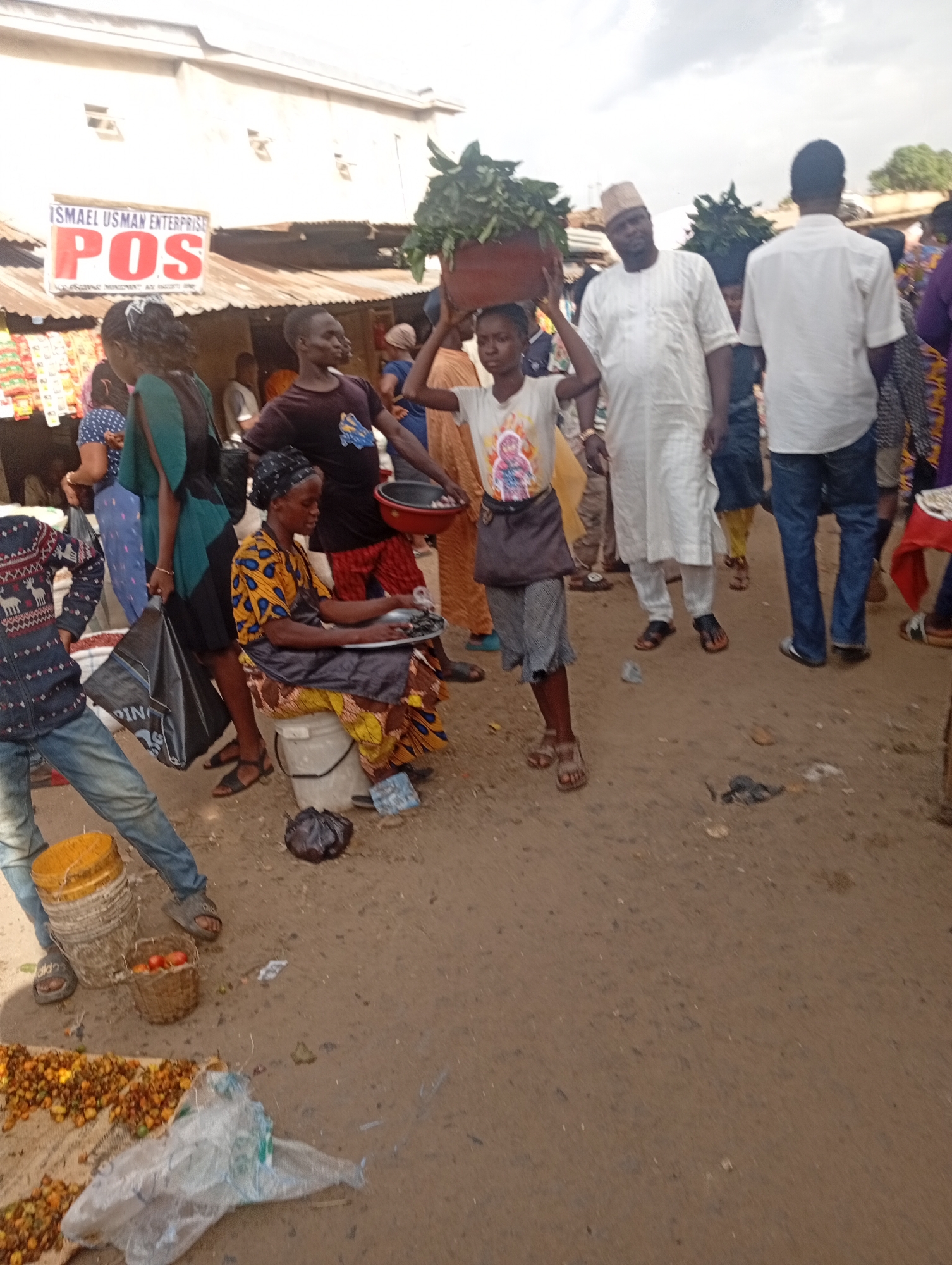 The image depicts a bustling outdoor market scene in Rafin Par, Nigeria, under an overcast sky. A young girl, with dark hair and wearing a light-colored t-shirt with a graphic and a dark skirt, is prominently featured in the center, carrying a large pile of leafy green vegetables on her head. She appears to be walking. To her left, a woman wearing a yellow and black patterned dress is seated on a white bucket, holding a plate with fish. A black headscarf covers her hair. A man in a white striped robe and a light-colored cap is walking to the right of the girl, looking towards the left. Another man in a white collared shirt and blue jeans is walking further to the right. Several other individuals are visible in the background, with some appearing to be vendors. In the foreground, a person in a blue sweater with a deer pattern and blue jeans is standing, facing away from the camera. A woven basket containing red tomatoes is near the person's feet, along with two buckets, one yellow and one white. Scattered on the ground are more food items, including what appear to be yellow fruits and vegetables, as well as discarded plastic. A black plastic bag and a clear plastic bag are also on the ground. A sign in the background reads "ISMAEL USMAN ENTERPRISE POS." The ground is covered in dirt and sand, typical of a market environment.