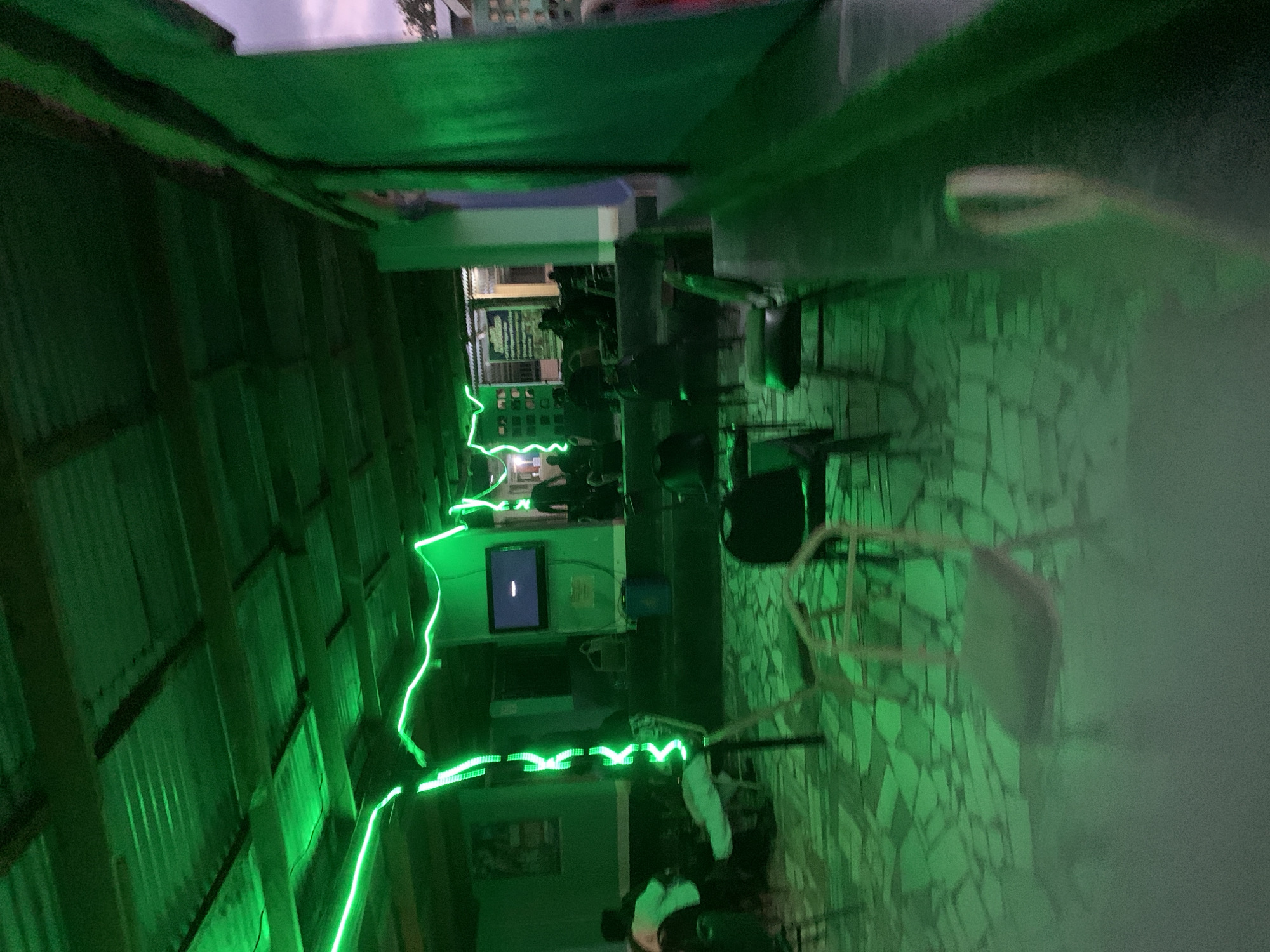 The image displays a semi-enclosed communal space located in the city of New Stadium, Nigeria, characterized by pervasive green artificial lighting. The ceiling features corrugated metal panels and visible support beams. Green LED string lights are strung in a wavy pattern across the ceiling and vertically down a central support, casting a dominant green hue over the entire area. The floor is covered with irregularly shaped tiles, also reflecting the green light.
Numerous simple, metal-framed chairs are distributed throughout the space, some occupied and others empty. In the mid-ground, a wall-mounted television screen displays a dark image with a prominent horizontal white line. A blue cylindrical bucket or container rests on the floor directly beneath the television. A poster is affixed to the far left wall. Approximately 5-10 individuals are present, primarily seated in various sections of the room. One person in the mid-ground is seated, wearing a light-colored shirt and trousers, with their legs extended. The overall illumination is dim, with the green light being the primary source. To the right and in the background, a purplish sky is visible, indicating either dusk or night, along with glimpses of adjacent outdoor structures.