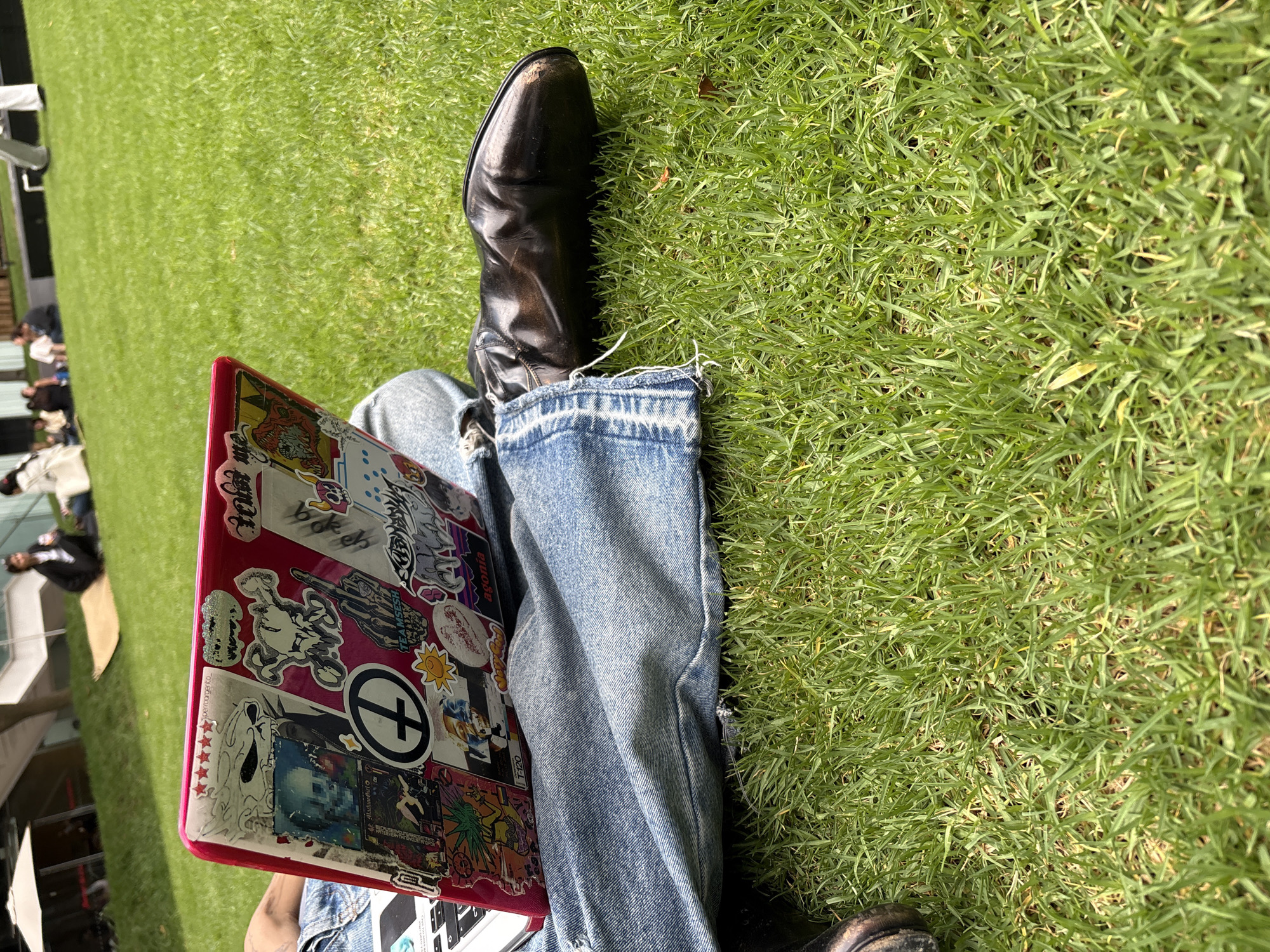 A ground-level shot captures a person sitting on a grassy area. The person is wearing light blue jeans and black leather boots. On their lap rests a pink laptop covered in stickers. The background is primarily green grass. In the distance, blurred figures of people can be seen, along with some structures that appear to be part of a tent or building. No specific information is available to determine if this photo was taken in Mexico City.