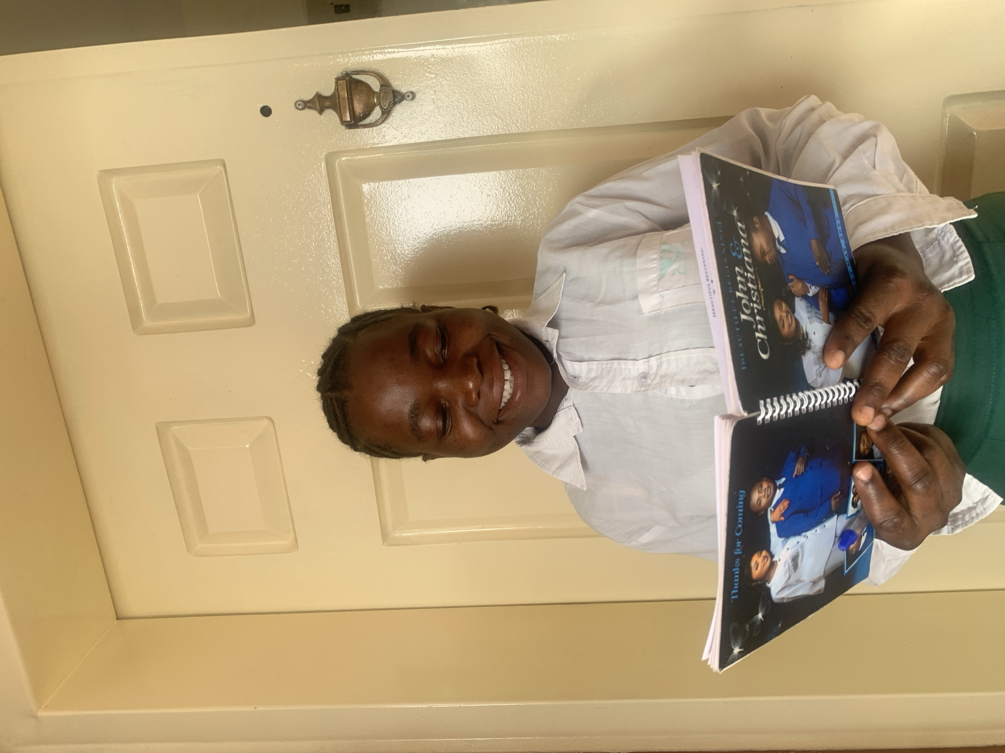 A woman smiles, holding open a spiral-bound booklet. The text on the cover reads "Beautiful Beginning! John & Christiana". The booklet features a photograph of a couple, the man in a blue suit and the woman in a white dress. Another page reads "Thanks for Coming". The woman holding the booklet wears a light-colored button-down shirt and dark green bottoms. She stands in front of a light-colored door with a metal knocker. The image was taken in Jos, Nigeria.