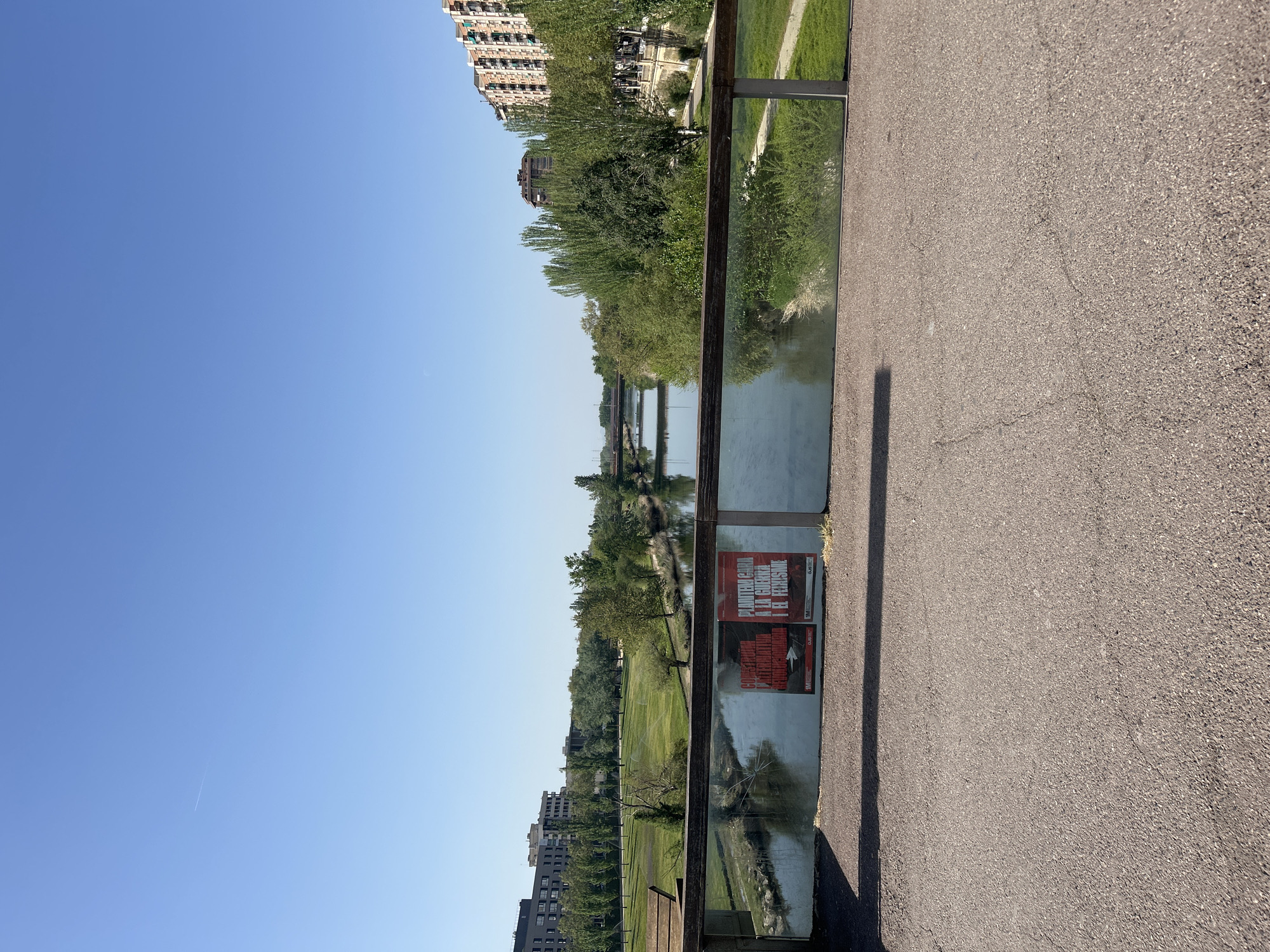 Here is a description of the image:
The image shows a view of the Segre River in Lleida, Spain, from a raised walkway or bridge. A clear blue sky takes up the left side of the image. The river runs through a park-like area with green grass and trees visible on both sides. Buildings are seen in the distance at both ends of the river stretch. A pedestrian walkway runs along the right side of the image with a glass barrier. Two posters are affixed to the glass barrier. The walkway surface appears to be made of asphalt.