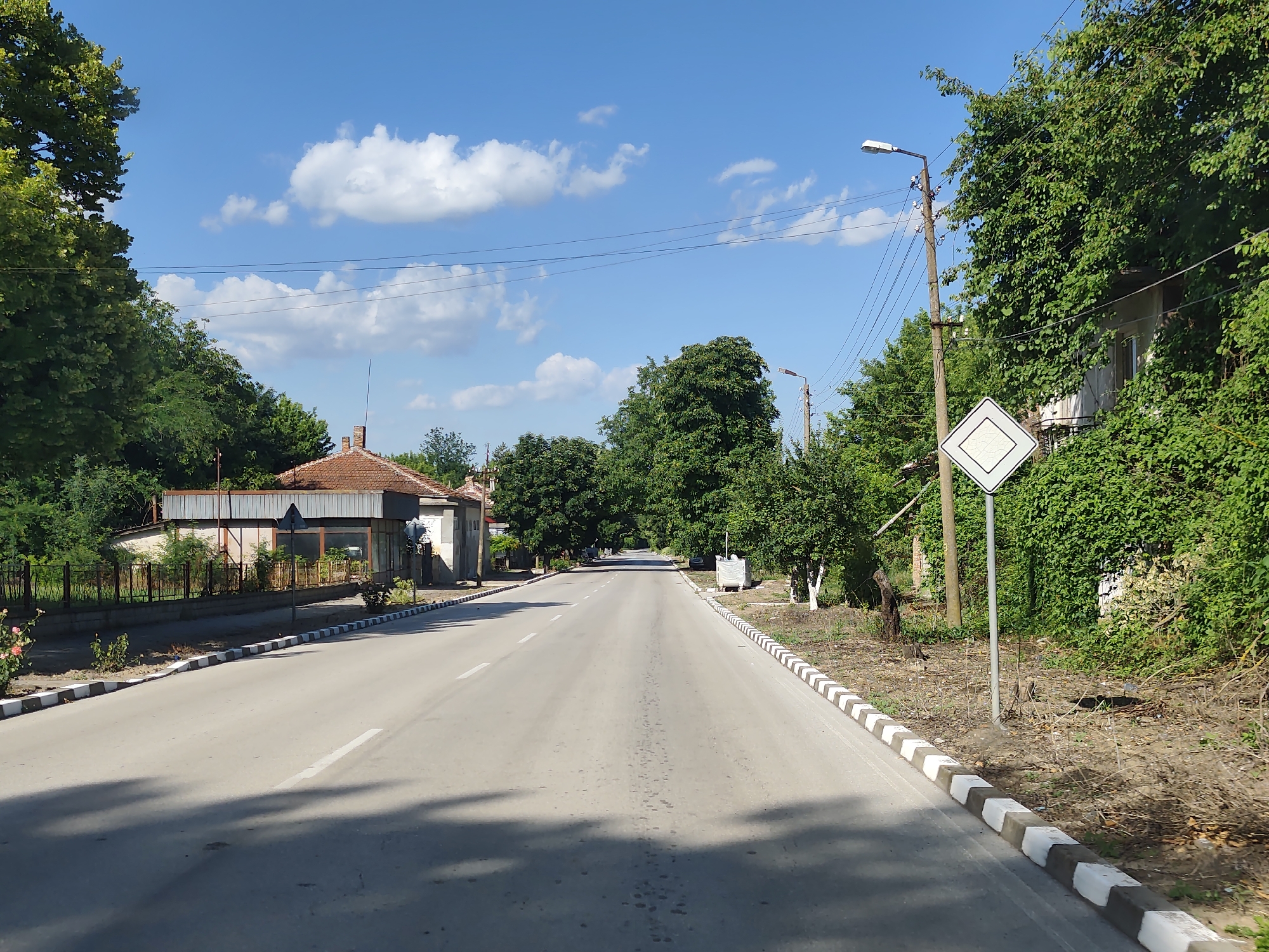The image depicts a straight, paved road in the village of Strezherovo, Bulgaria. The road has a white dashed line marking its center and is bordered by alternating black and white curbs. On the left side of the road, there are buildings with red tile roofs, fences, and lush green trees. On the right side, more trees and vegetation line the road, with a white diamond-shaped road sign on a pole and a street light visible. The sky is blue with scattered white clouds. The scene appears to be captured during daylight, likely in the summer, given the foliage. There are no people or vehicles visible in the image. The overall impression is of a quiet, rural road in a Bulgarian village.