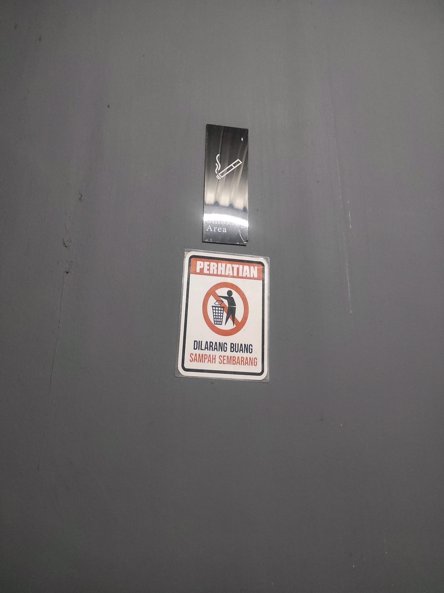 The image shows two signs on a grey wall. The top sign is a rectangular "No Smoking" sign with an image of a cigarette and smoke. Below this is a smaller sign that reads "Area". The second sign is also rectangular and has a red border. It contains the word "PERHATIAN" at the top, followed by a pictogram of a person with a trash can and a diagonal line through it, indicating "No Littering". Below the pictogram, it reads "DILARANG BUANG SAMPAH SEMBARANG", which translates to "Do not litter". This image was taken in South Jakarta, Indonesia.