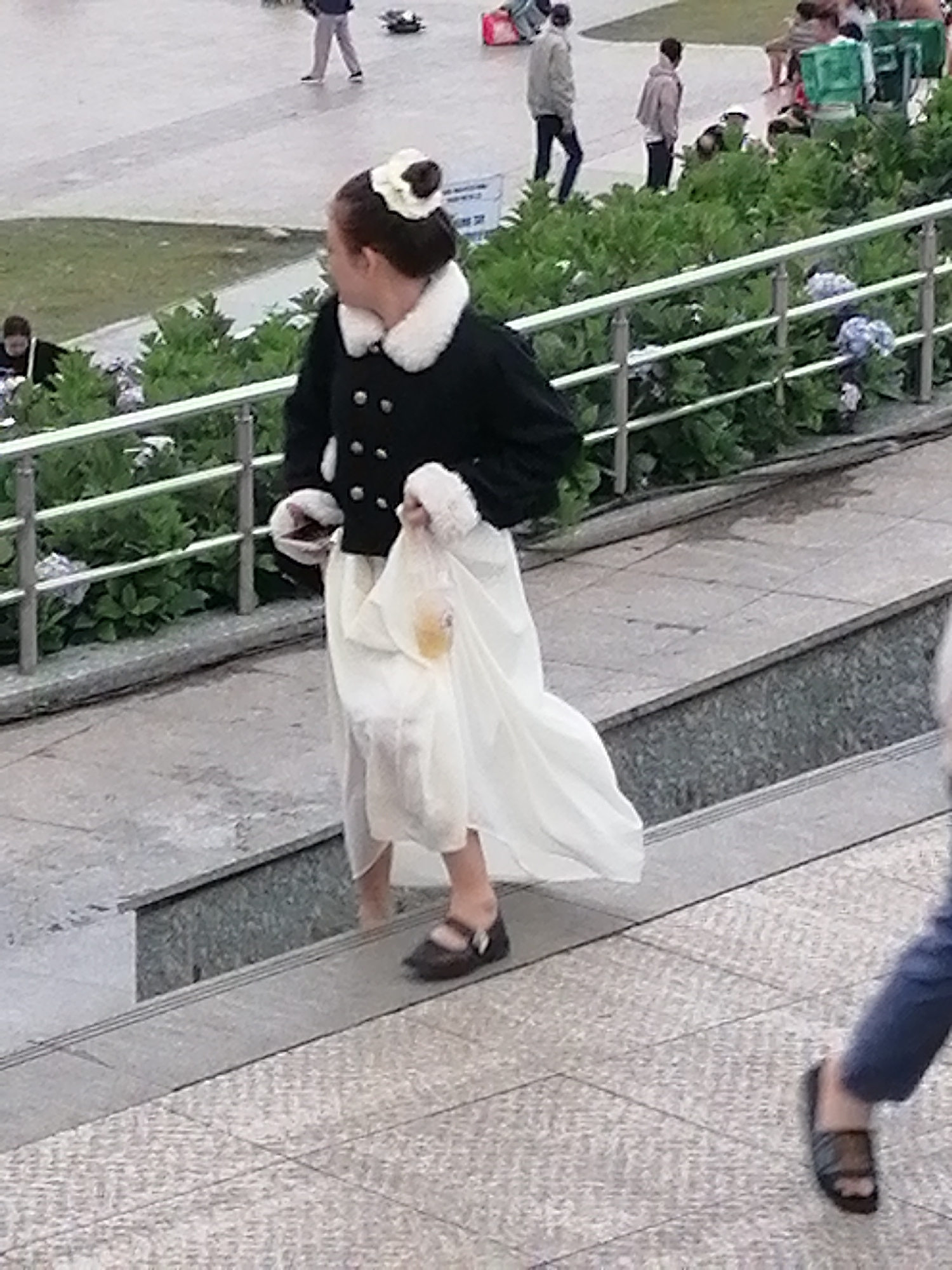 A woman wearing a white and black outfit with a fluffy collar and white trim is walking down stone steps outdoors. She has a white bow in her hair and is holding a clear plastic bag with a yellow object inside. The steps are surrounded by green foliage and a silver metal railing. Other people are visible in the background, some standing and others walking. The scene appears to be in an urban environment, possibly a public park or a street in Da Lat, Vietnam, characterized by its paved walkways, greenery, and stone structures.