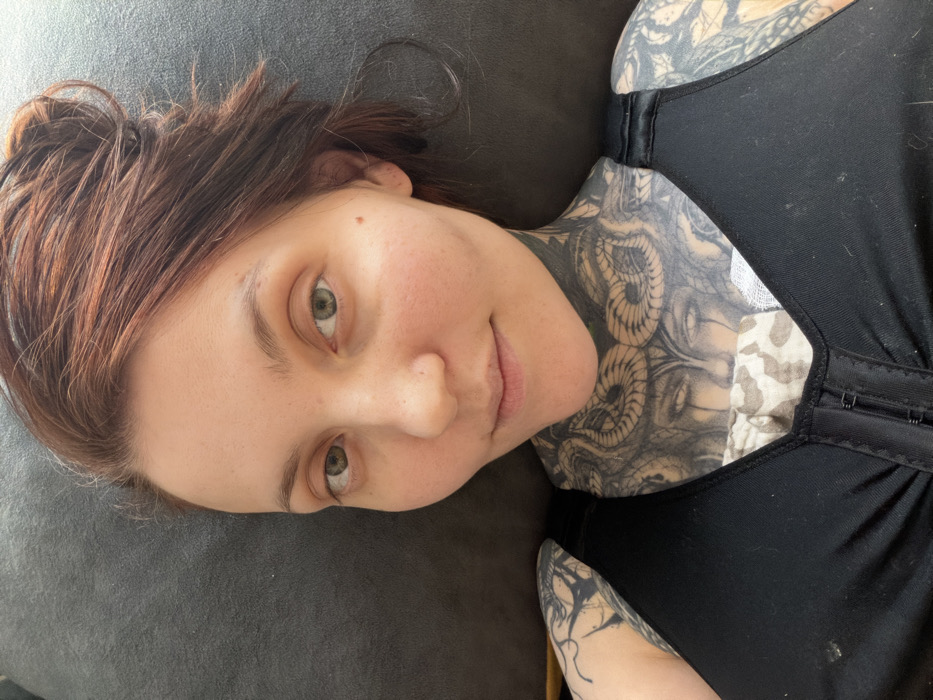 An overhead photograph depicts an adult female lying supine, looking directly at the camera. She has reddish-brown hair styled loosely and green eyes. Her face shows no obvious makeup. She is wearing a black compression-style garment or bra with a front hook-and-eye closure. Visible beneath this garment, near the sternum, is a light-colored fabric with a faint pattern. Extensive black and grey tattoos cover her neck, shoulders, and arms. The neck tattoo features intertwining snakes and a face. She is positioned against a solid, dark grey or black textured surface, likely fabric. The precise location within the city of Chaikovsky, Russia, cannot be determined from the visual information presented in the image.