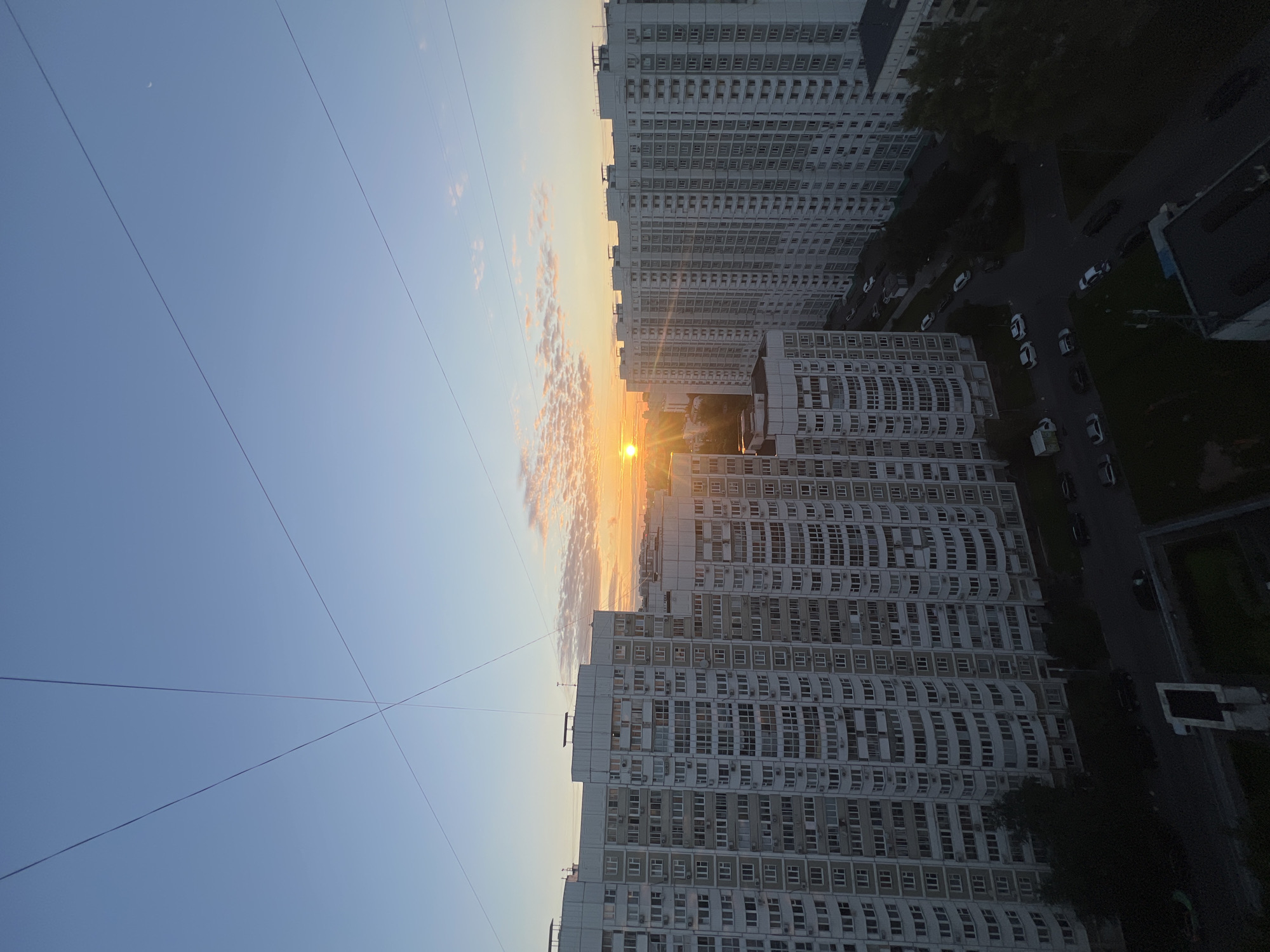 This is an eye-level, wide-angle outdoor shot of a residential area in Moscow, Russia, during sunrise. The sky is a gradient of pale blue and soft orange, with scattered, wispy clouds. Several power lines crisscross the sky. In the foreground, towering, multi-story apartment buildings dominate the scene. These buildings are characterized by their light-colored facades and numerous windows, many of which are crescent-shaped, a common architectural feature in Russian apartment blocks. The buildings are densely packed, with narrow streets and parking areas visible between them. Several cars are parked along the roads. The sun is low on the horizon, casting a warm glow over the scene and creating a bright lens flare. The overall impression is one of an urban, early morning landscape.