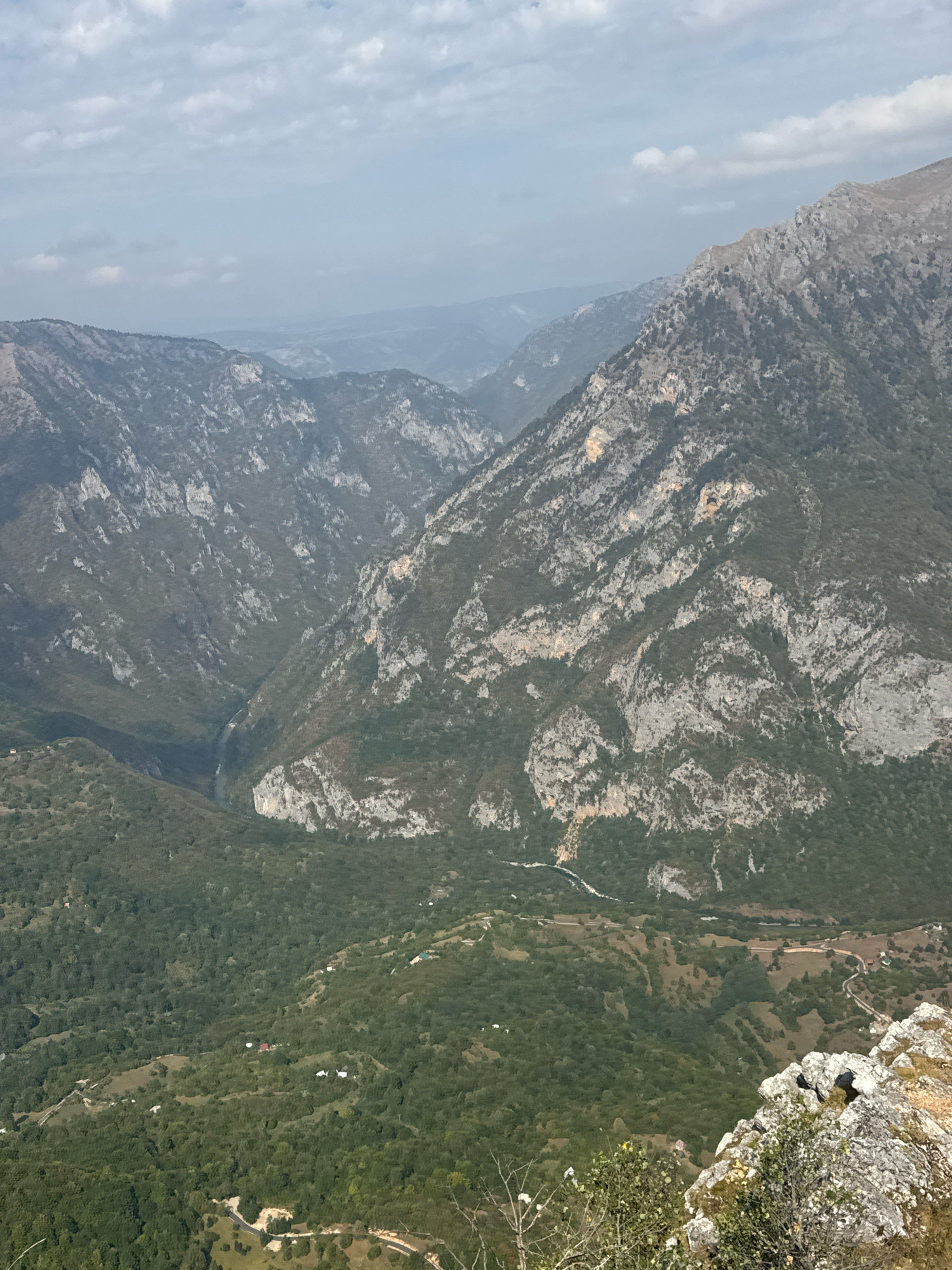 The image depicts a panoramic view of a vast mountain range, likely in the vicinity of Žabljak, Montenegro, known for its dramatic landscapes and canyons. The foreground is dominated by rugged, rocky terrain with sparse vegetation, suggesting a high vantage point. Beyond this, the landscape unfolds into a series of steep, verdant mountains and deep valleys. The mountains are characterized by their rocky, uneven surfaces, interspersed with patches of dark green forests that cling to the slopes. A winding river or road can be faintly observed in the distance, cutting through the valley floor and adding a sense of scale to the immense natural beauty. The sky is partly cloudy, with soft, diffused sunlight casting a gentle glow over the scene, hinting at either early morning or late afternoon. The overall impression is one of remote, untamed wilderness.