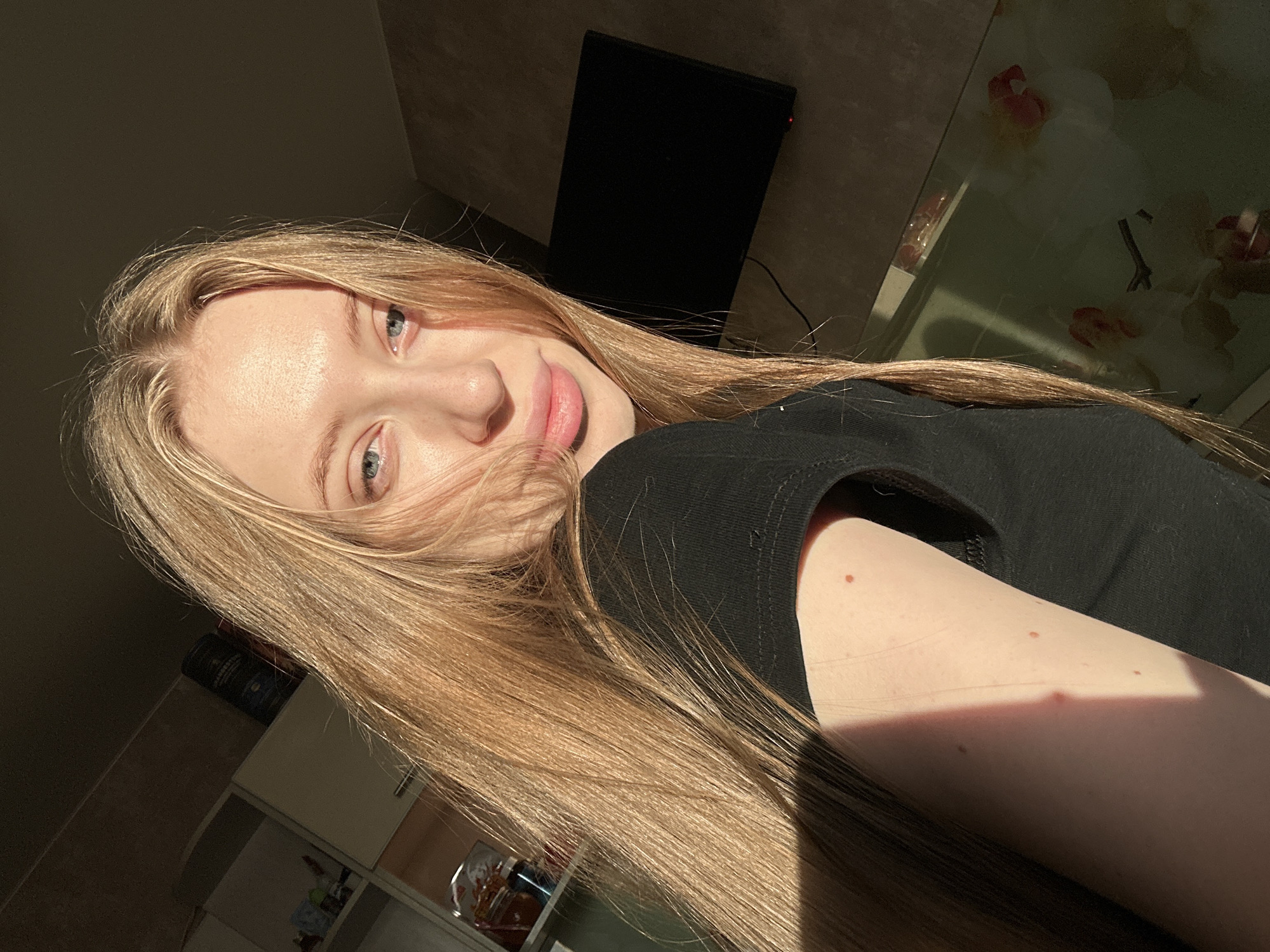 A young woman with long, straight, light blonde hair and blue eyes is captured in an indoor setting, likely a residential kitchen or dining area in Chaikovsky, Russia. She is positioned facing the camera, with her head slightly tilted, and her lips slightly parted. Strong direct sunlight illuminates the right side of her face and hair (viewer's left), casting shadows on the opposite side. She is wearing a black short-sleeved top. In the background to her left, white kitchen cabinets are visible, some with glass doors revealing items inside. A dark, cylindrical container rests on top of one cabinet. To her right, a black flat-screen television is mounted on a dark, textured wall. Below the television, a reflective surface, possibly a tabletop, shows indistinct reflections.